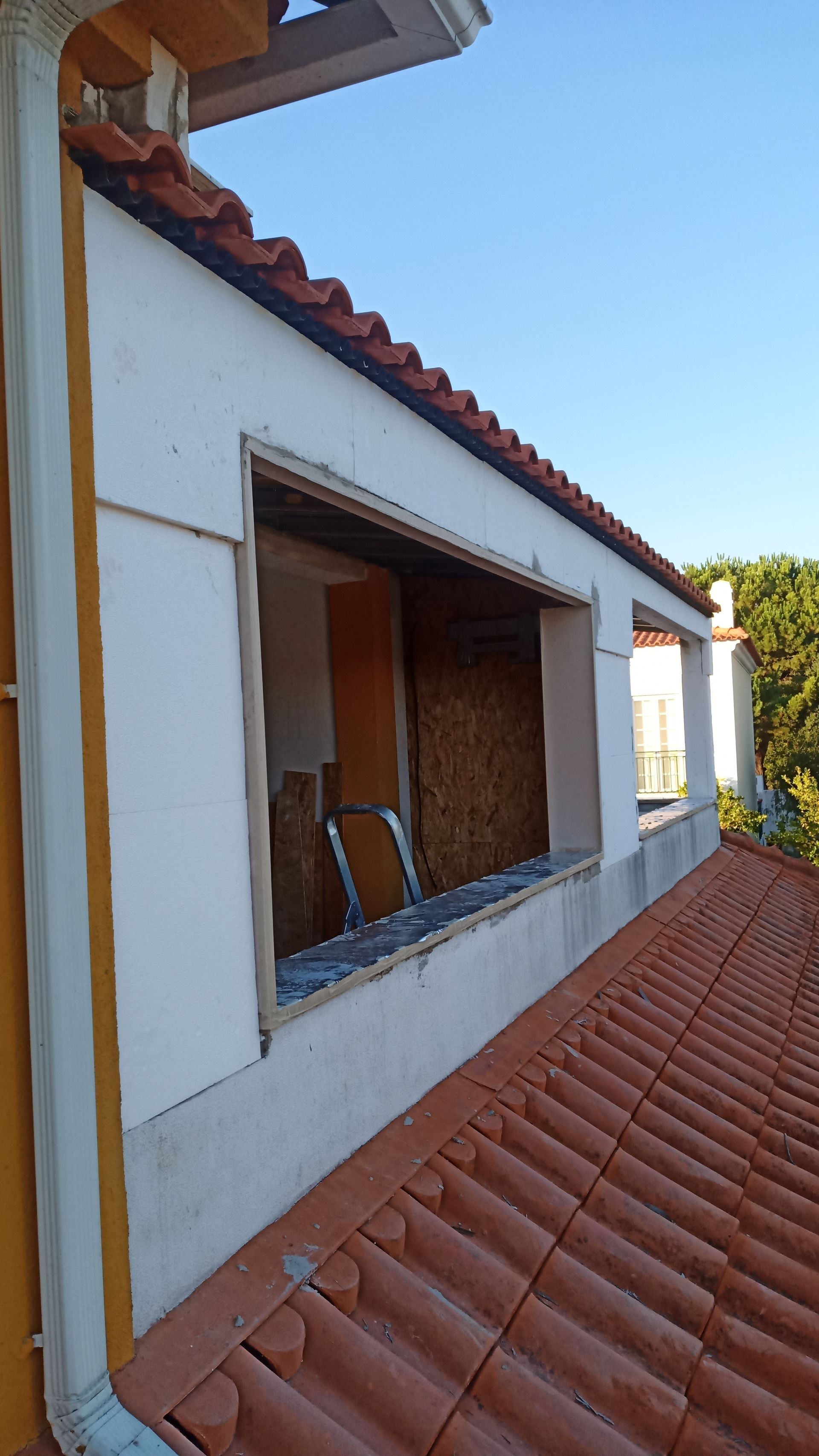 Caixilho de janela sendo construído em um telhado de telhas vermelhas, com tábuas cobrindo a abertura e paredes brancas.