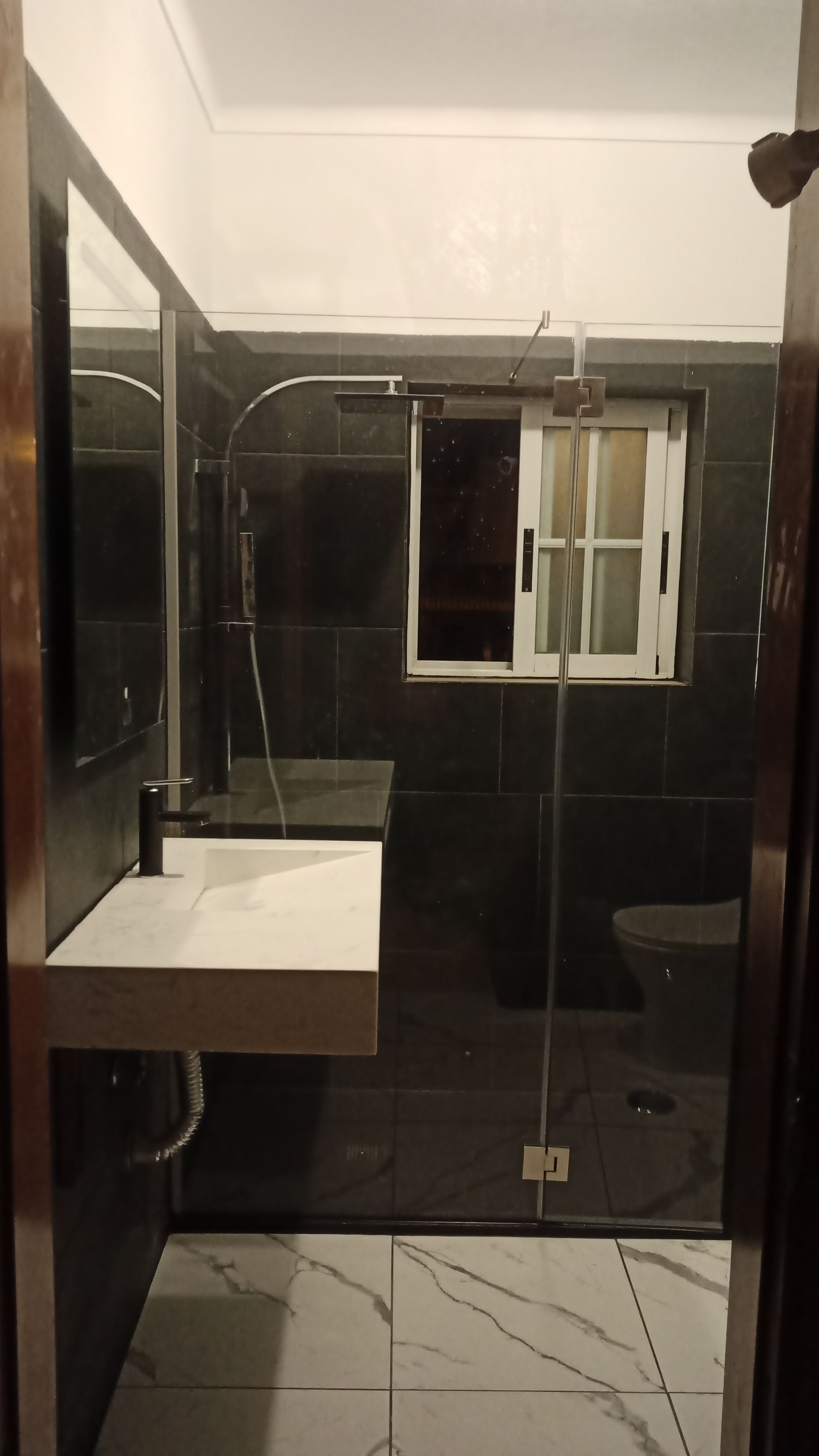 Banheiro moderno com paredes revestidas de azulejos pretos, pia branca e box de vidro. Piso de mármore branco.