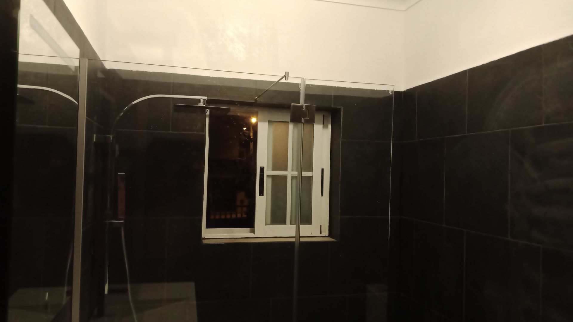 Box de chuveiro com azulejos escuros, paredes de vidro, janela ao fundo e chuveiro cromado.