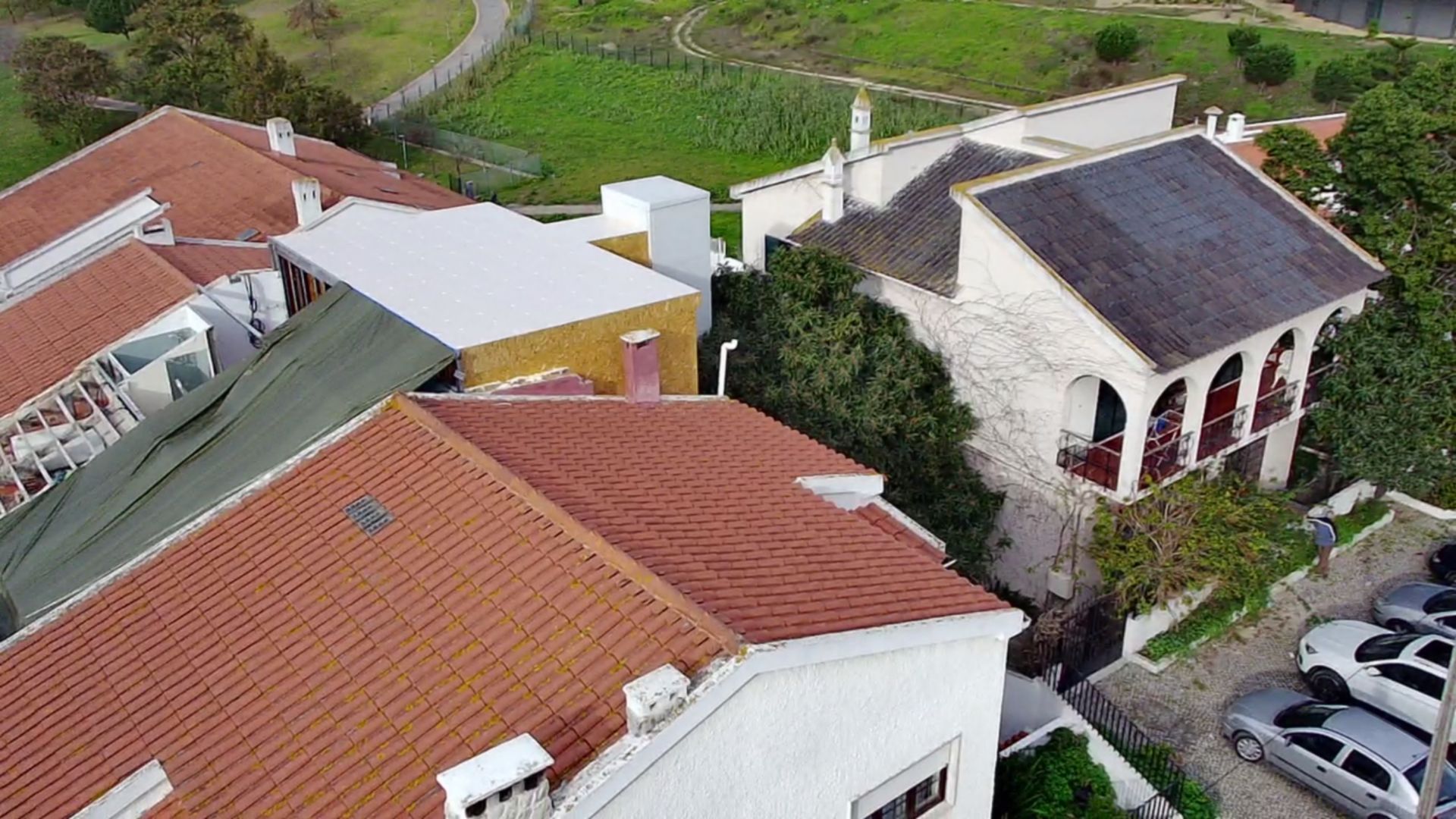 Prédios com telhados de telha laranja, um deles com varanda, cercados por vegetação e carros.