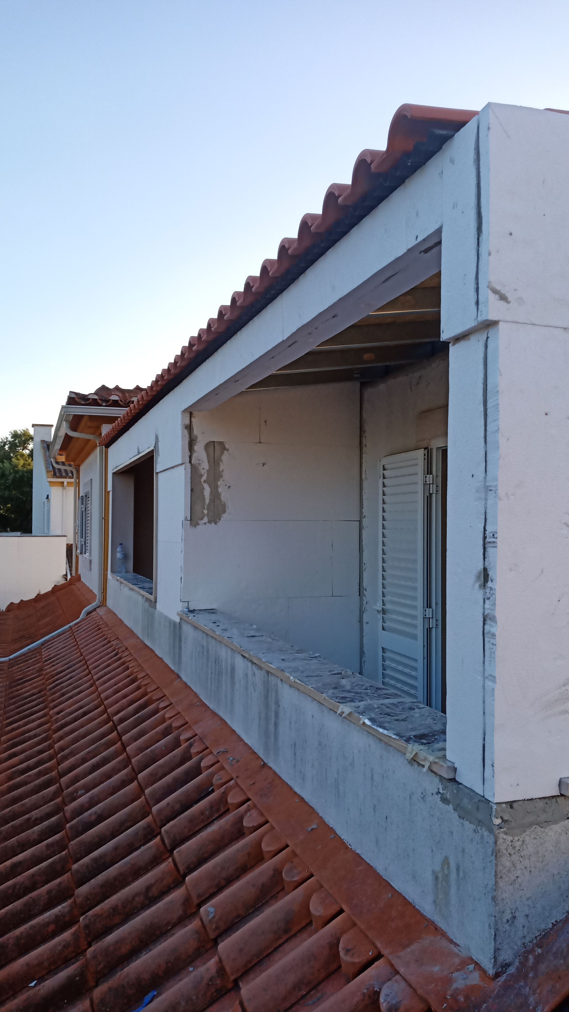 Vista do terraço de um prédio com telhado de telhas vermelhas, paredes brancas, janela aberta e céu azul.