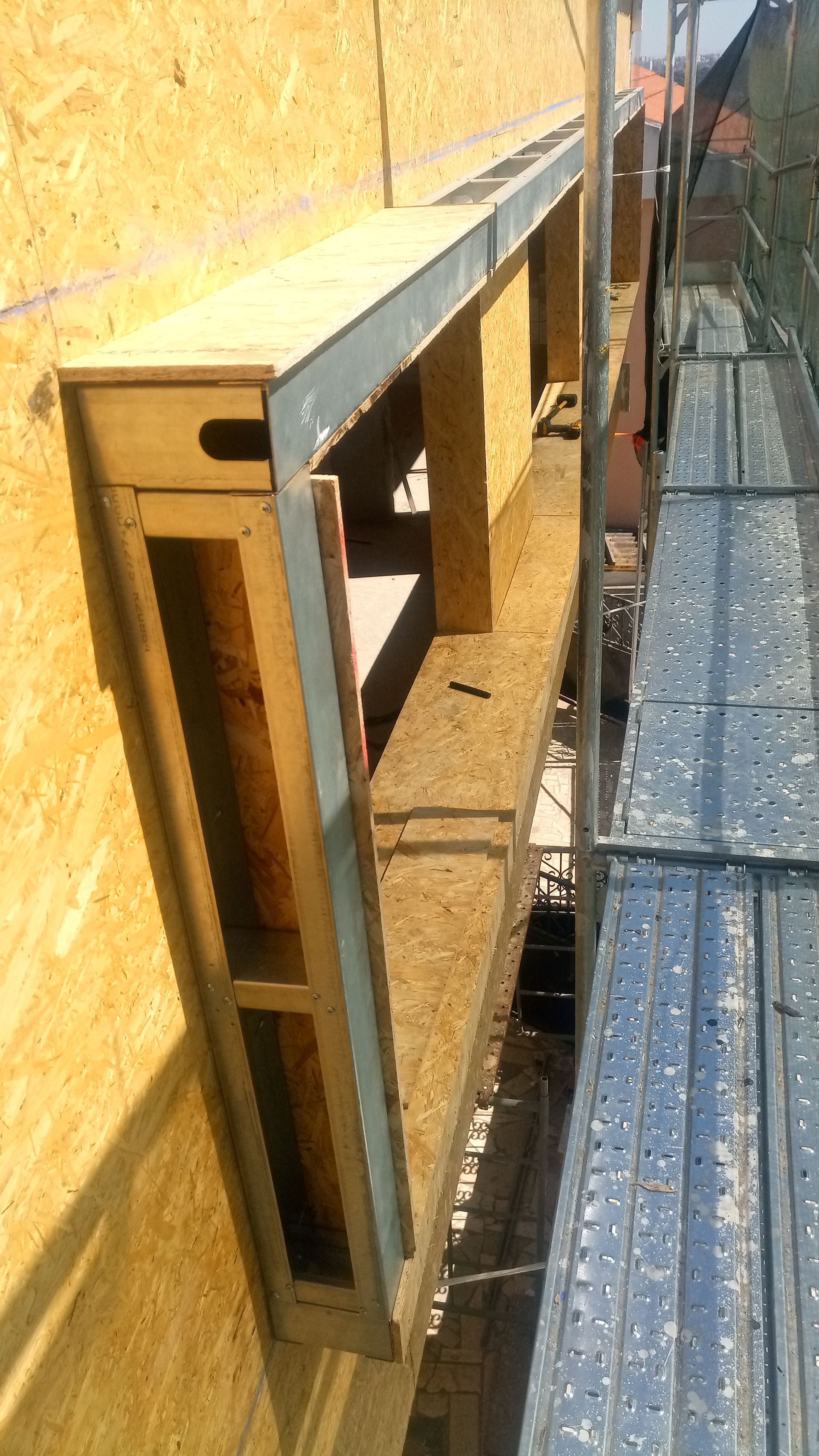 Estrutura de madeira em forma de caixa, montada na parte externa de um edifício, com andaimes próximos.