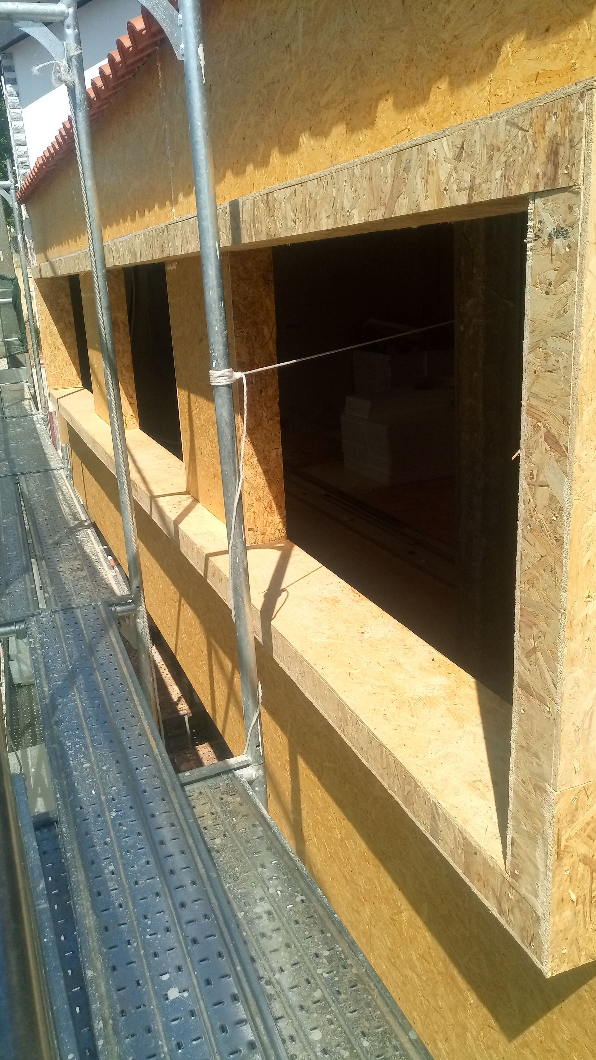 Parede externa em construção com caixilhos de janelas, madeira bege, andaimes e telhas vermelhas visíveis.