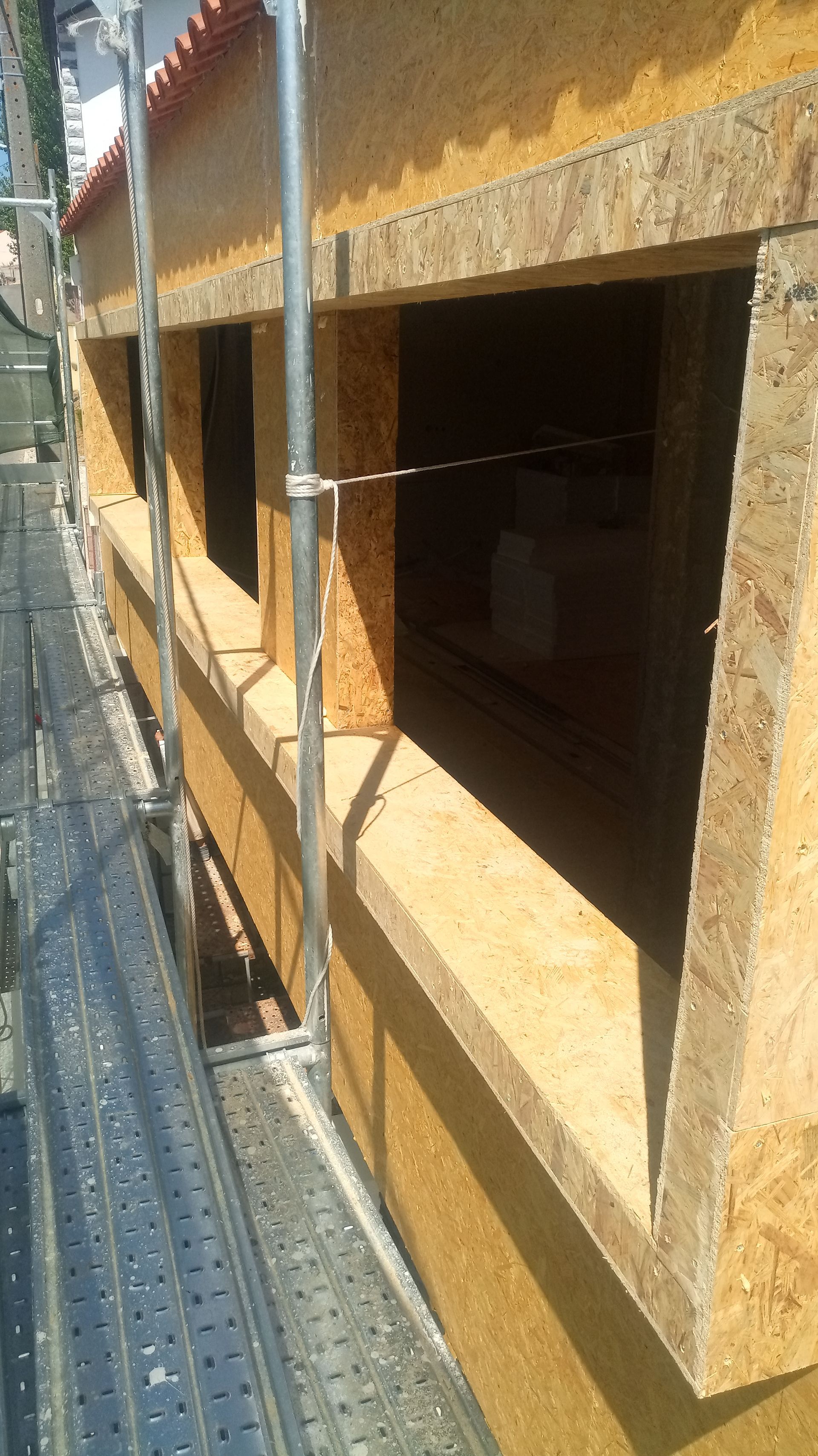 Parede externa de um edifício em construção, apresentando várias aberturas de janelas, andaimes e tábuas de madeira bege.