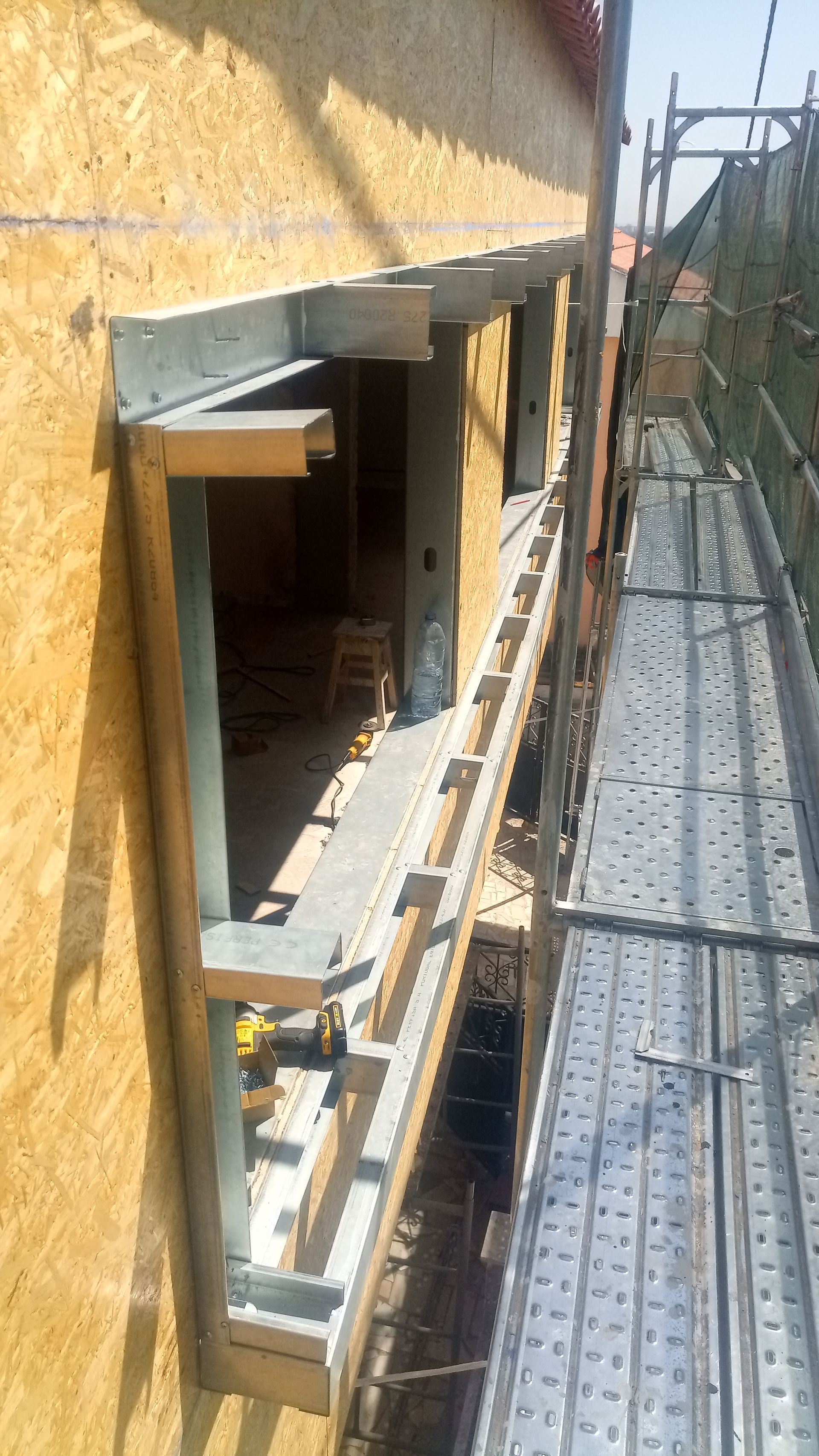 Exterior de um canteiro de obras com andaimes ao lado de uma parede inacabada. A moldura da janela é visível.