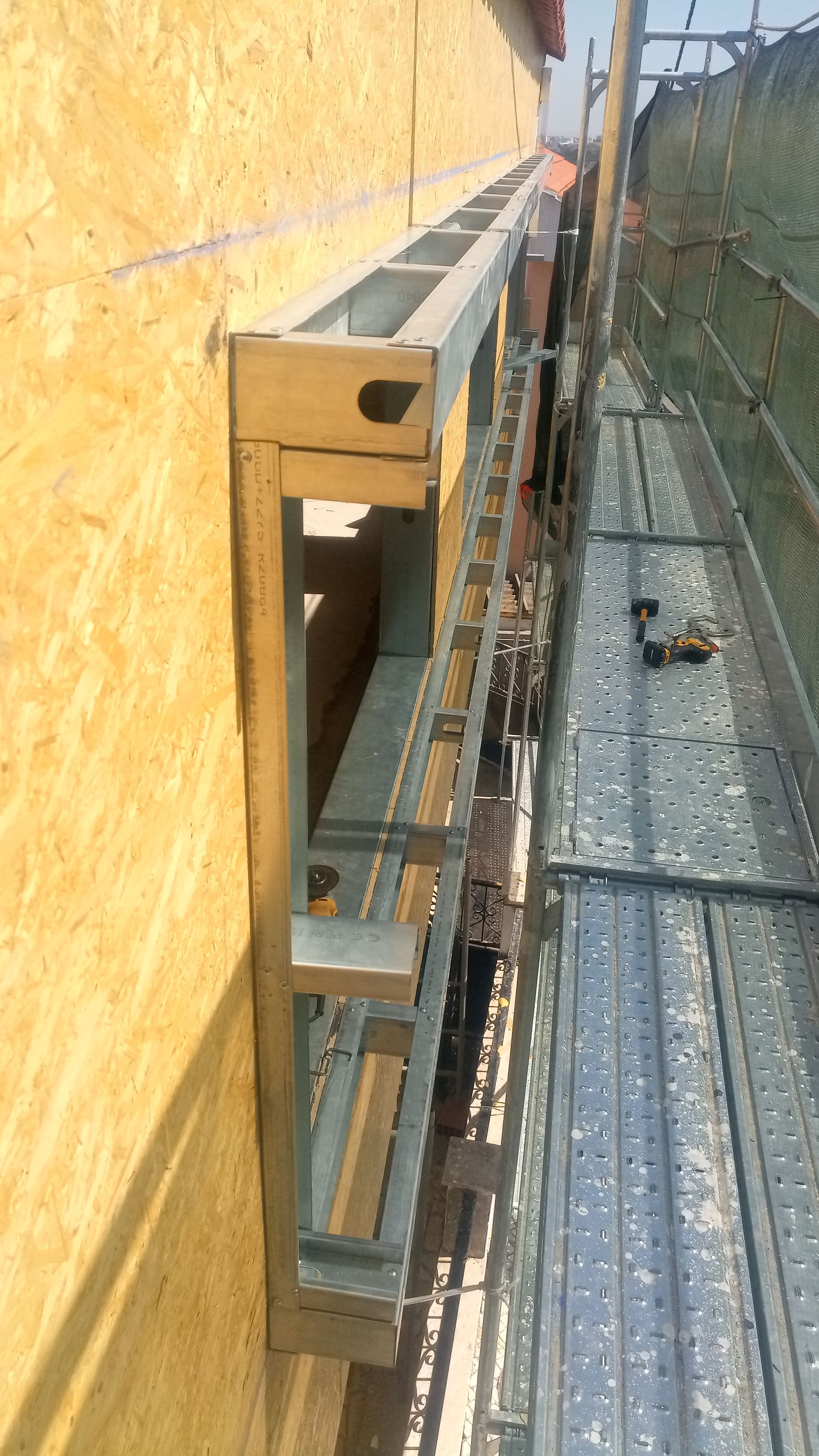 Estrutura metálica fixada a uma parede de OSB; andaimes próximos.