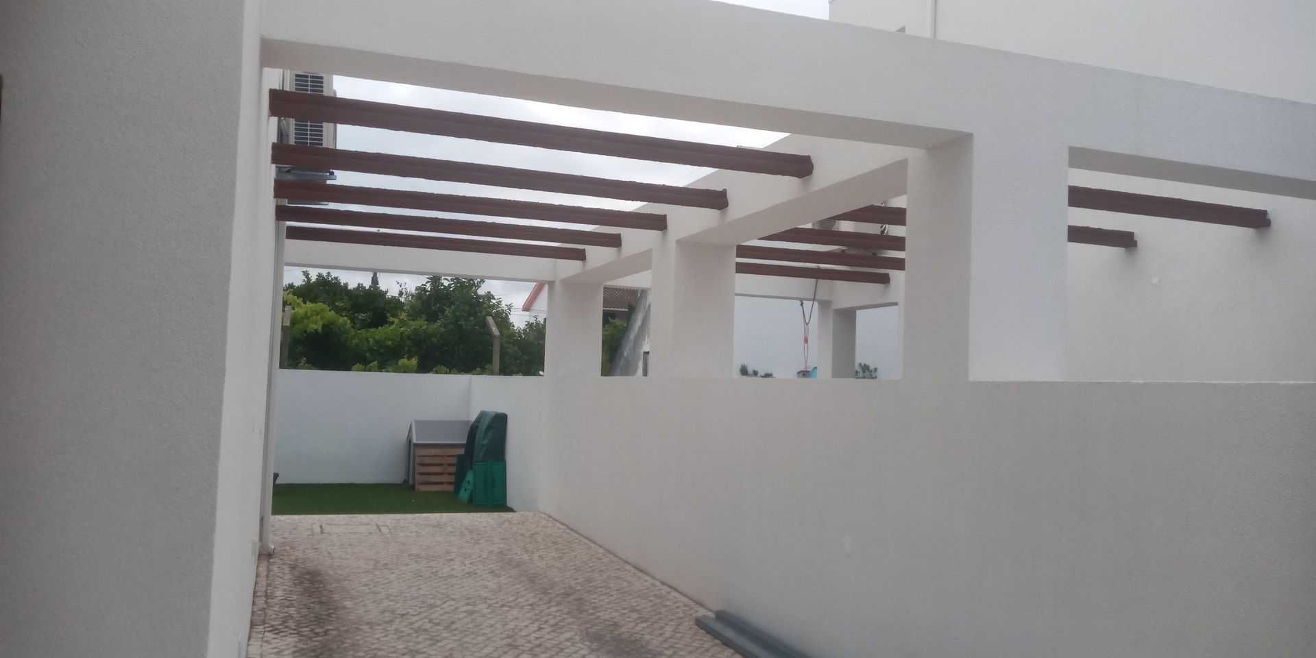 Pérgola branca com vigas marrons sobre uma entrada de garagem. Um muro branco se estende ao lado. Árvores são visíveis ao longe.