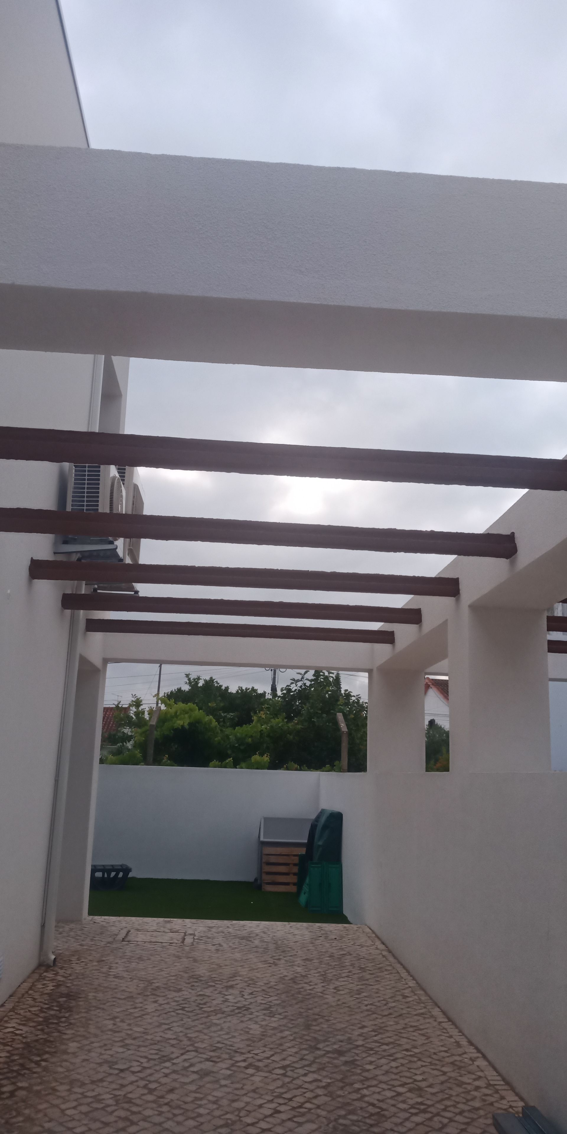 Área externa coberta com piso de seixos, paredes brancas e vigas de madeira. Folhagem verde visível ao fundo.