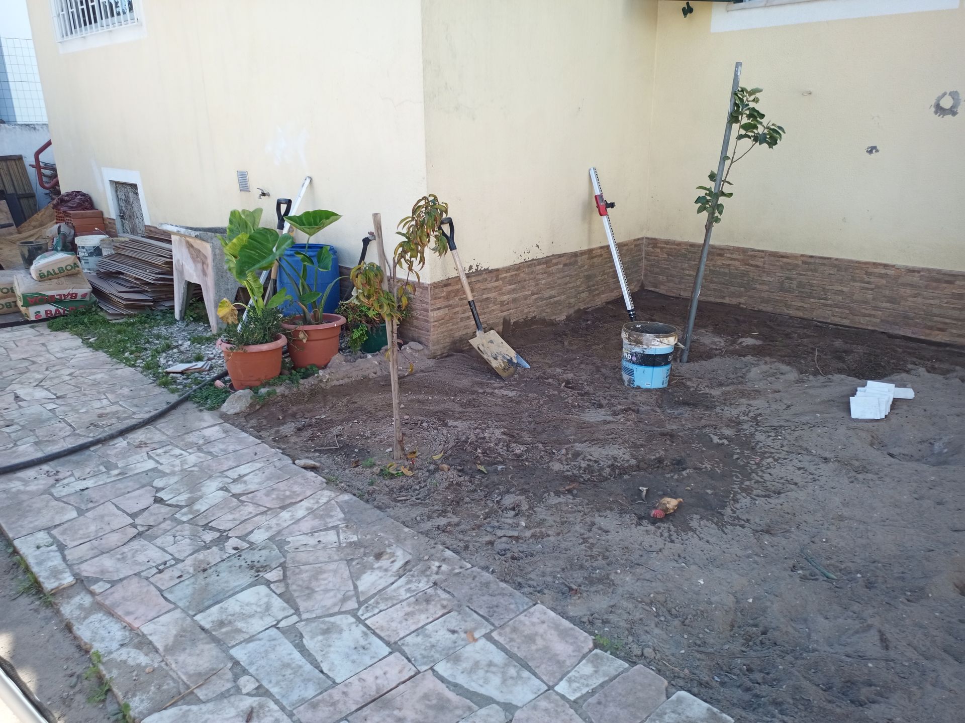 Quintal com caminho de pedra, plantas em vasos e terra pronta para plantio; mangueira e ferramentas disponíveis.