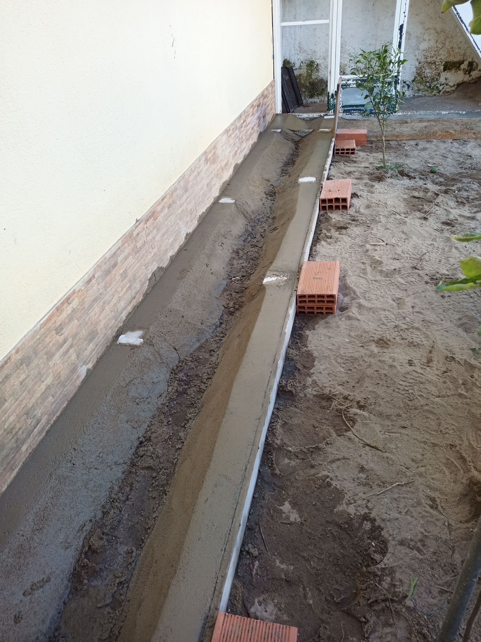 Construção de um caminho de concreto ao longo da parede de um edifício, com tijolos utilizados como marcadores.