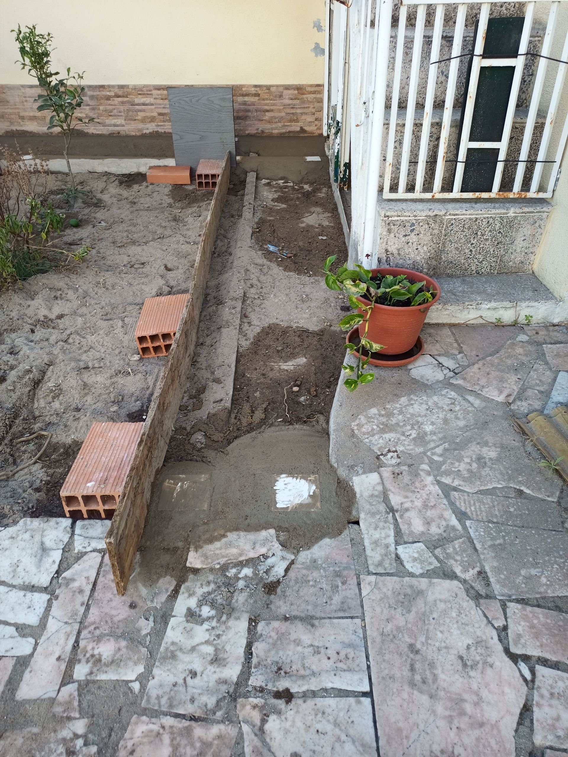 Canteiro de obras com calçada delimitada por tijolos e concreto, vaso de flores e porta.