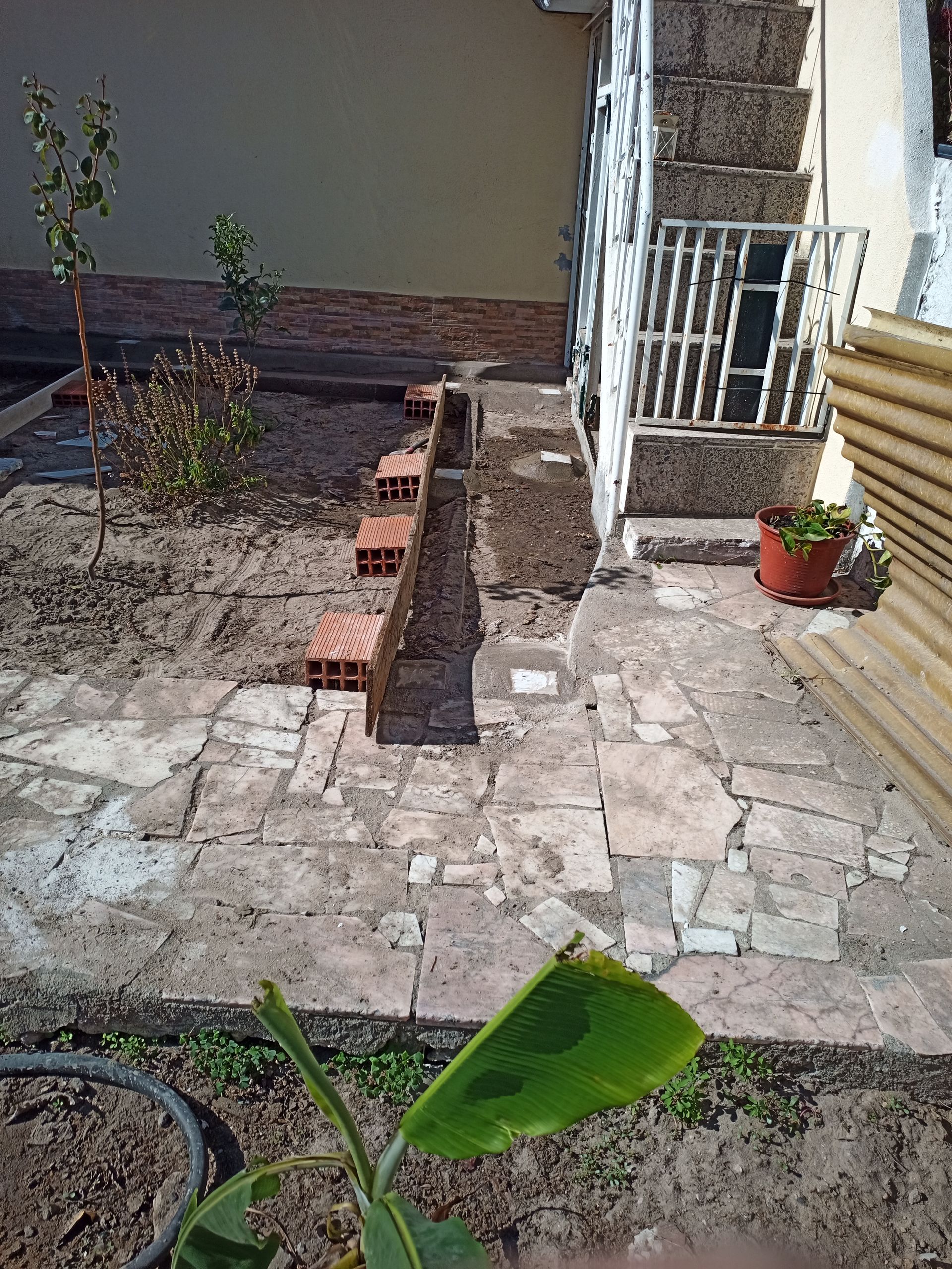 Quintal com caminho de tijolos, degraus e áreas ajardinadas; pátio marrom claro, construção com janela gradeada branca.