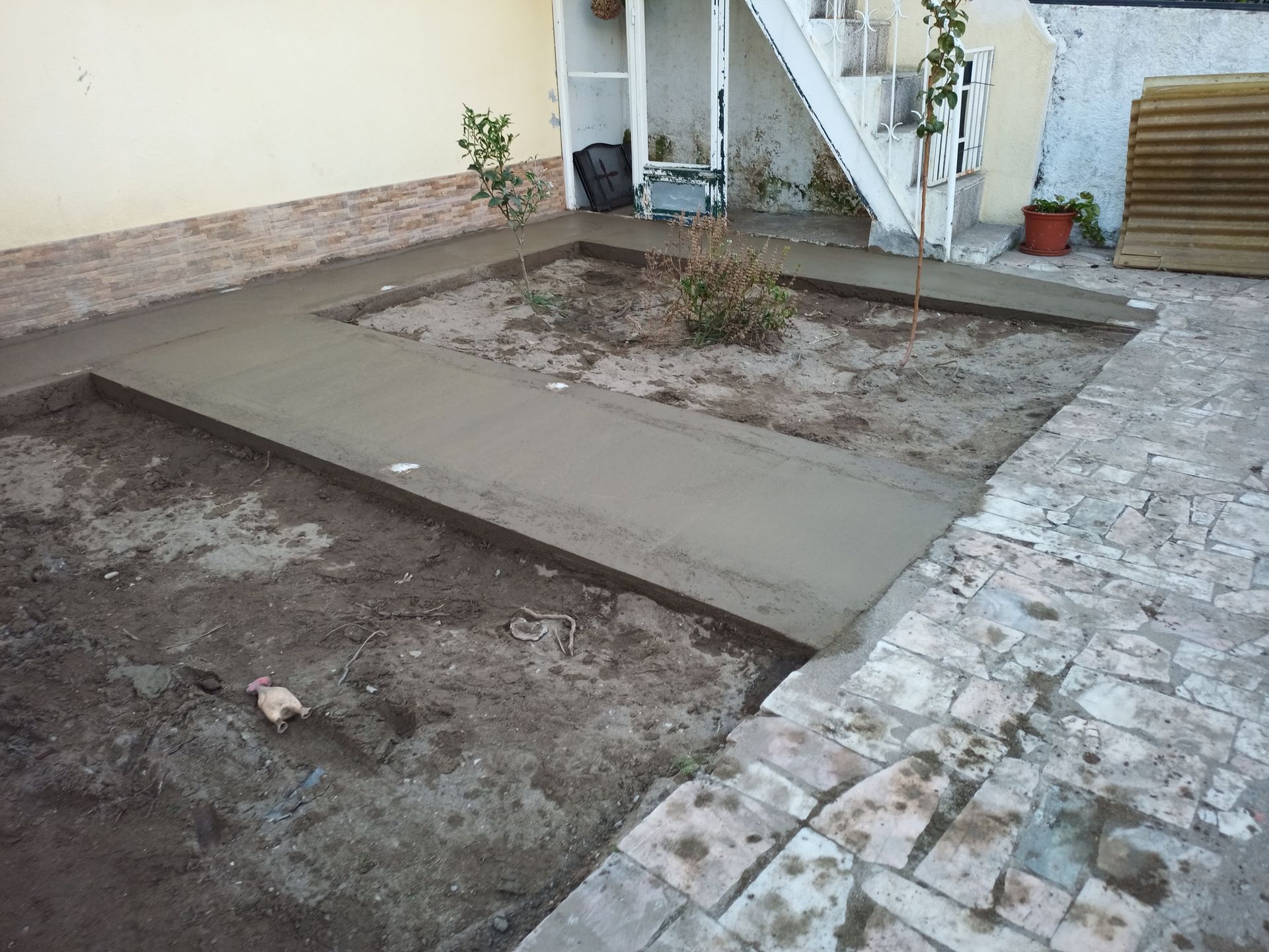 Calçada de concreto recém-construída em um quintal, cobrindo parcialmente a terra. Escadas e um prédio podem ser vistos ao fundo.