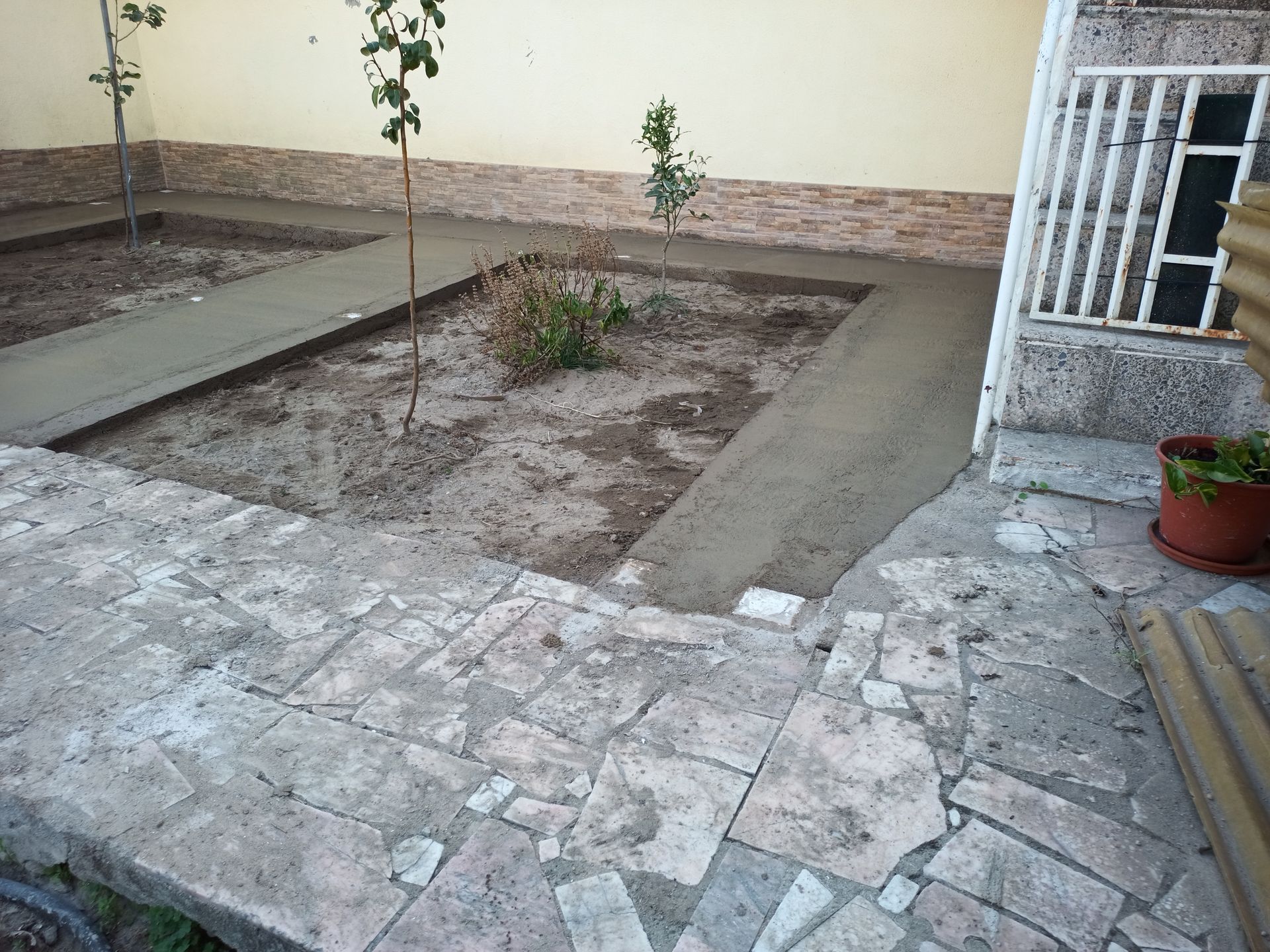 Caminhos de concreto e canteiros de jardim em um ambiente externo, ao lado de um prédio com um portão branco e um vaso de plantas.