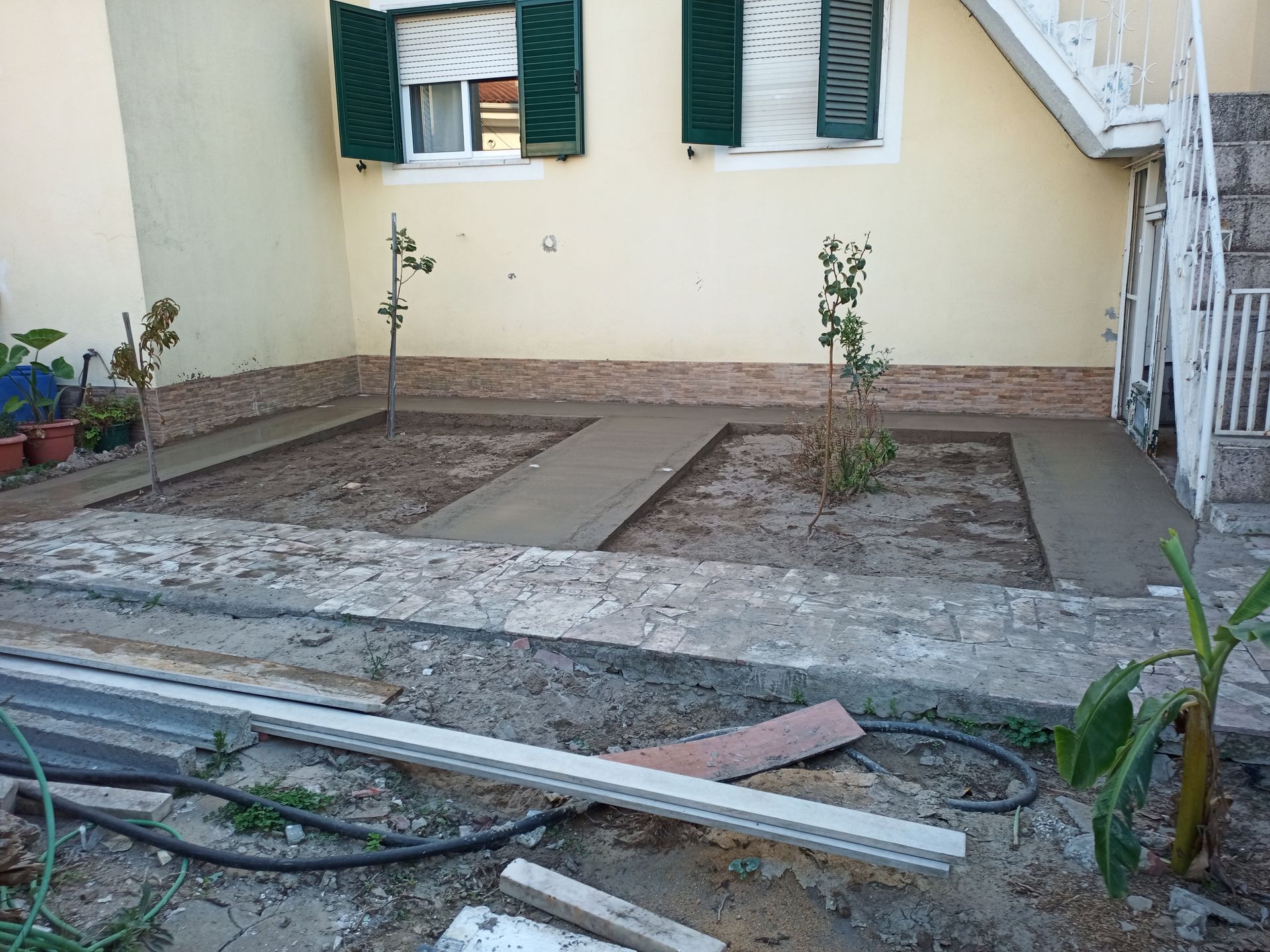 Jardim com caminhos de concreto e pequenas árvores. Casa com persianas verdes ao fundo.