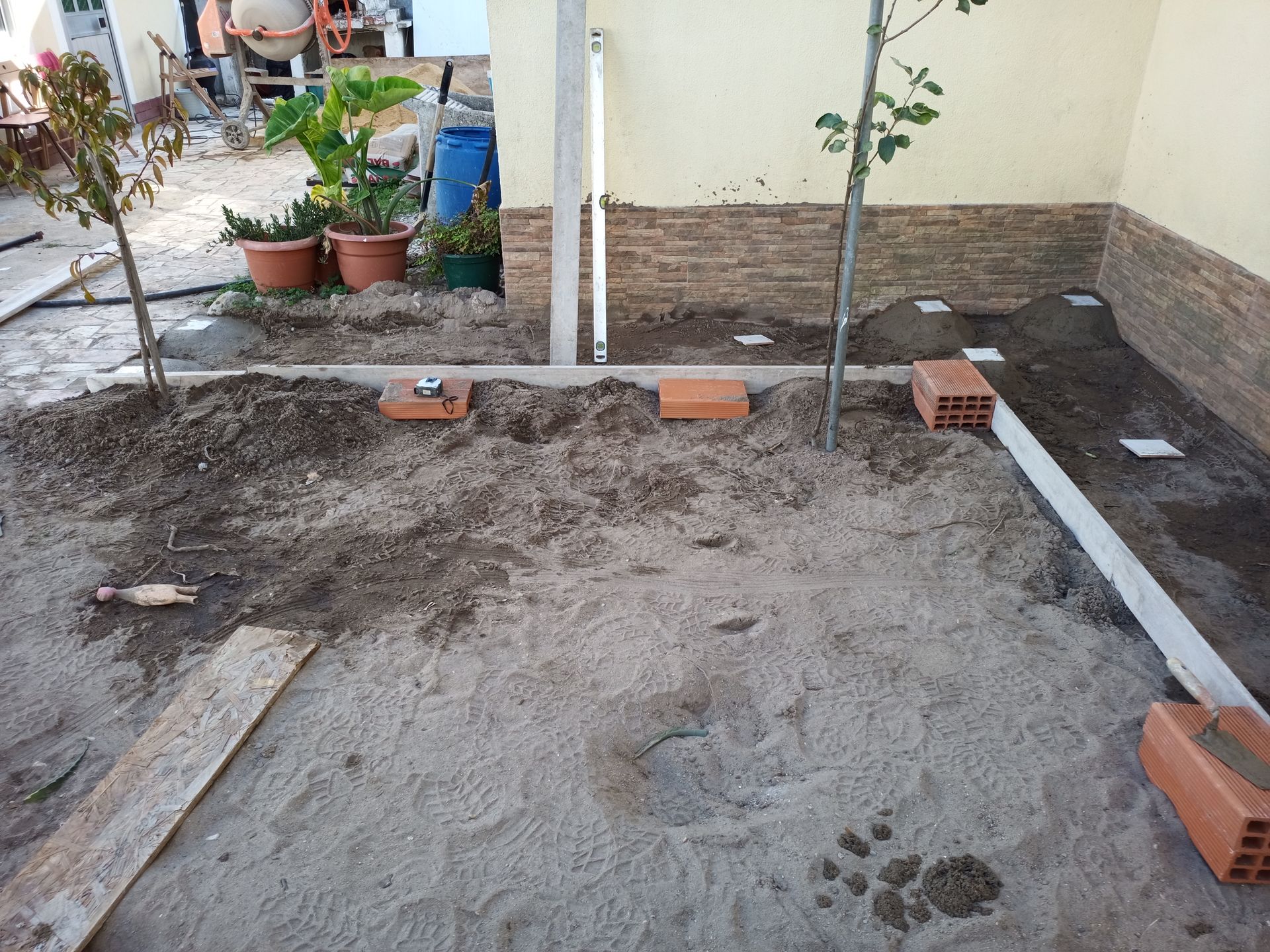 Uma área ajardinada com areia e terra, tijolos, plantas e uma parede parcialmente de cimento.