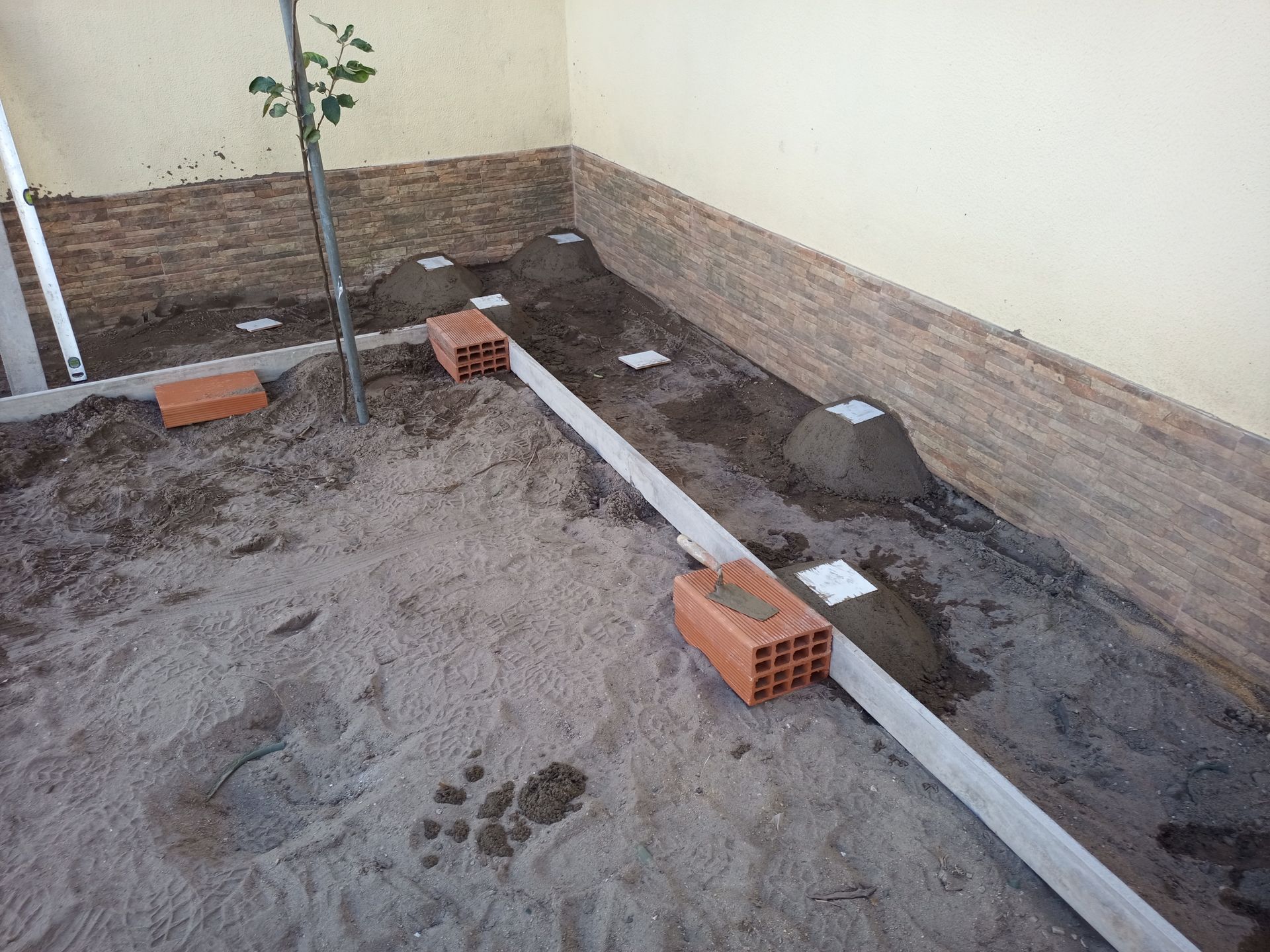 Um canteiro de obras com piso de areia, tijolos, blocos de concreto e tábuas de madeira já instalados; paredes ao fundo.