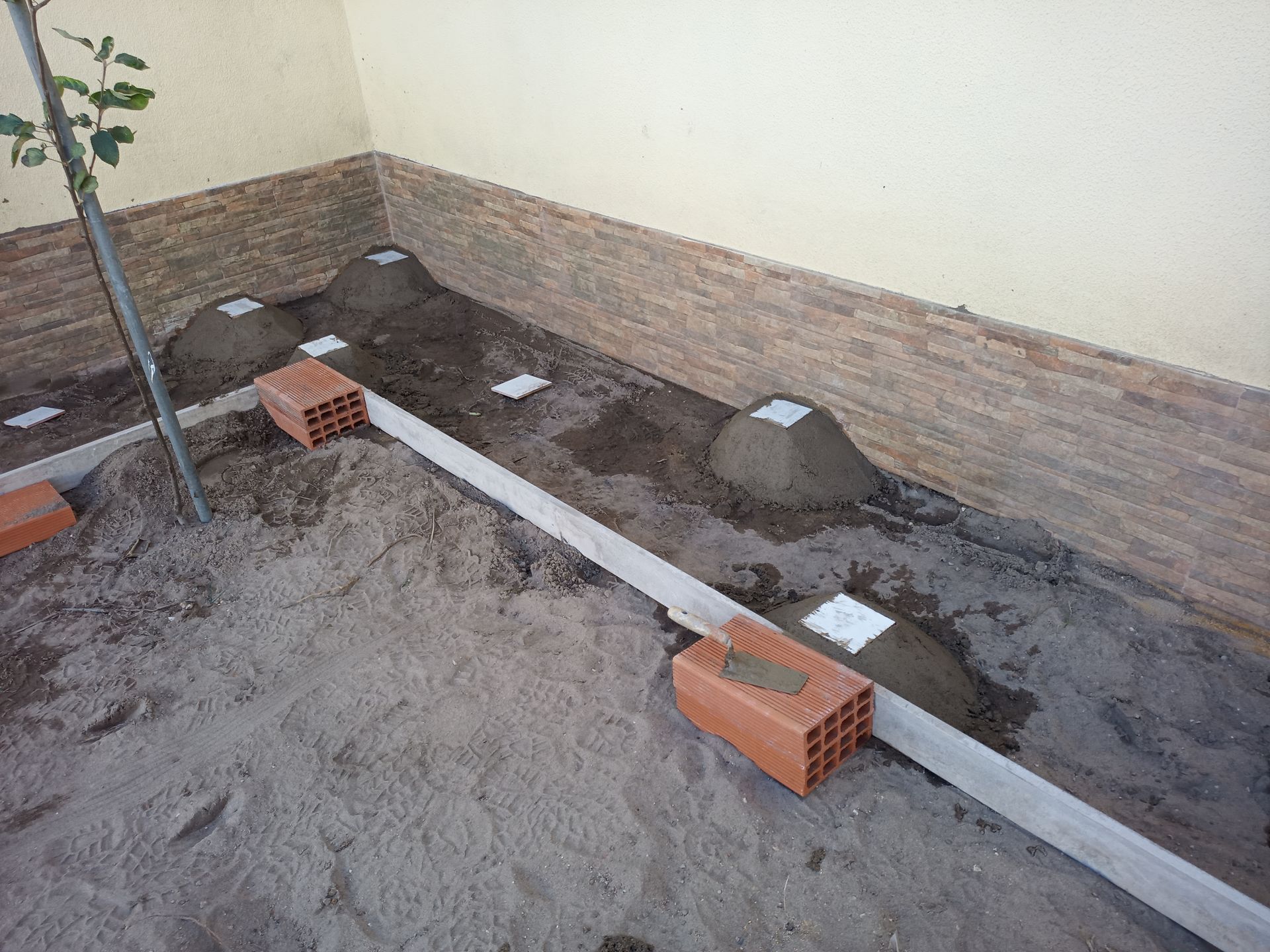 Canteiro de obras com areia, tijolos, montes de terra e uma tábua de madeira; próximo a uma parede.