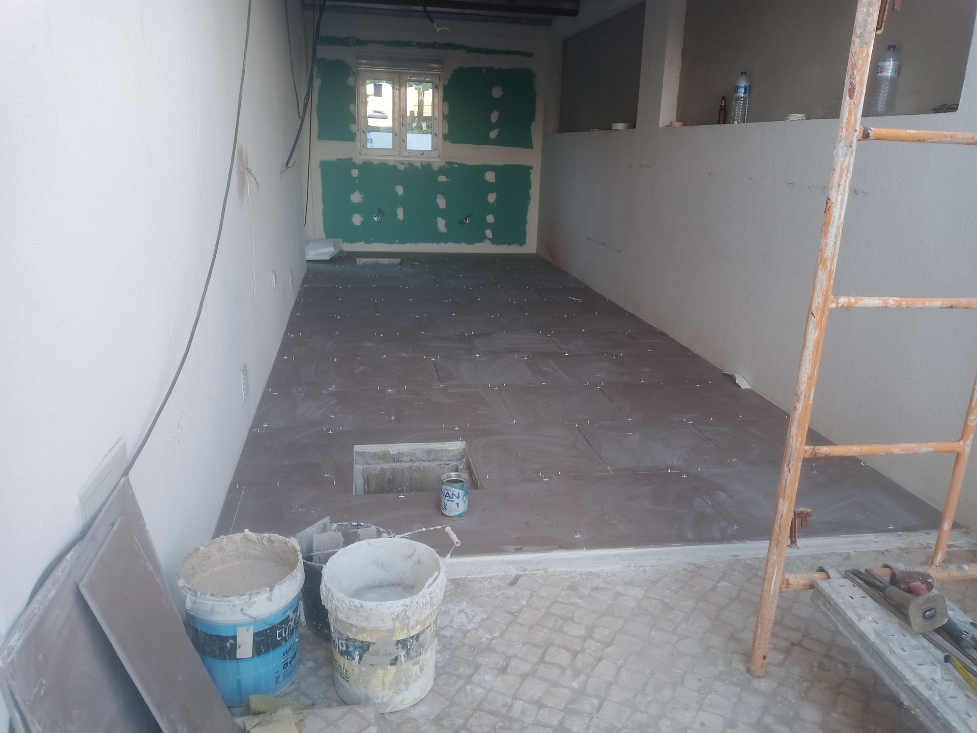 Quarto em construção, com piso parcialmente revestido com azulejos, vigas de parede expostas e baldes de tinta.