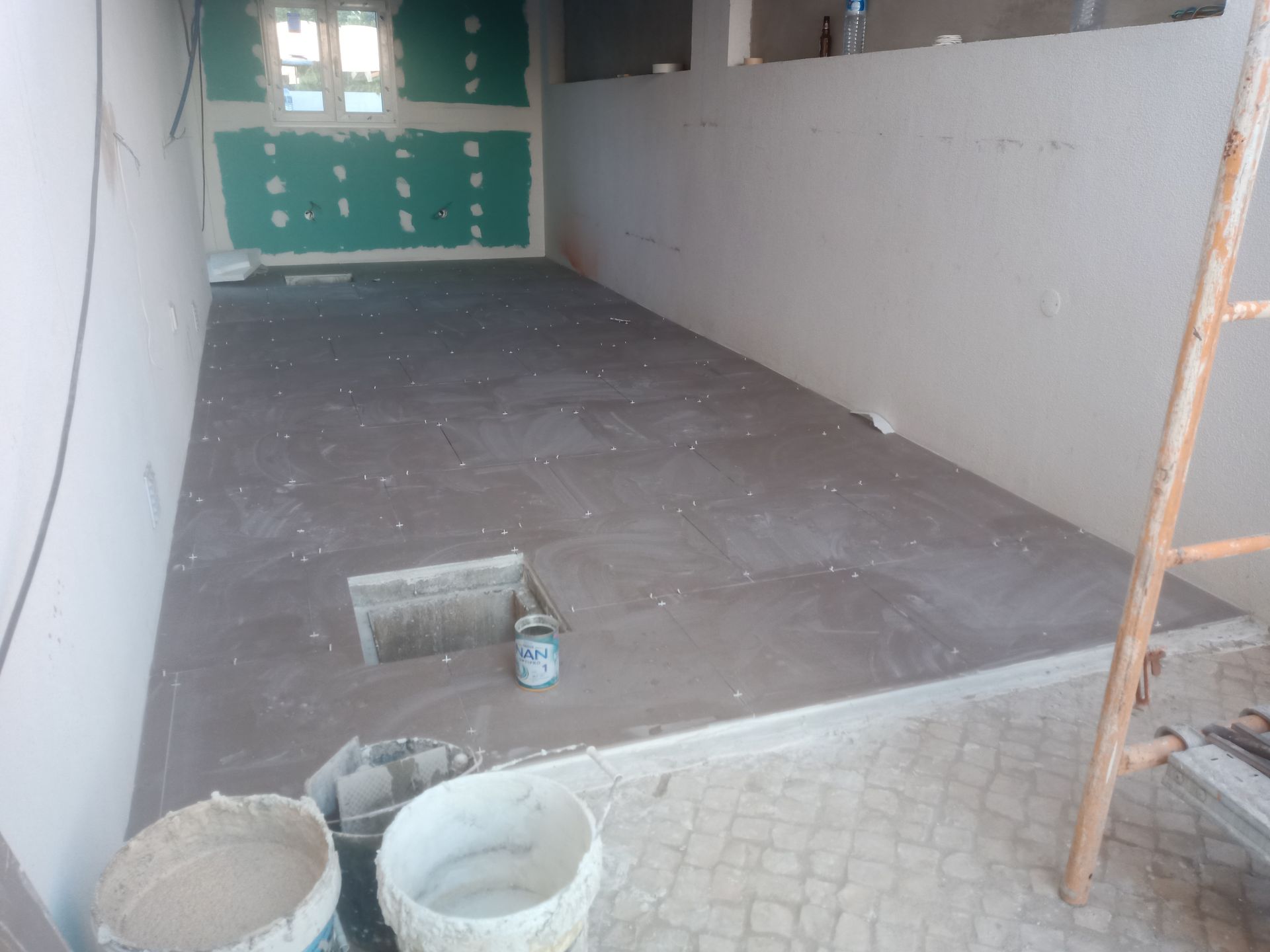 Canteiro de obras: Instalação de piso de azulejo, baldes de argamassa, paredes inacabadas, escada.