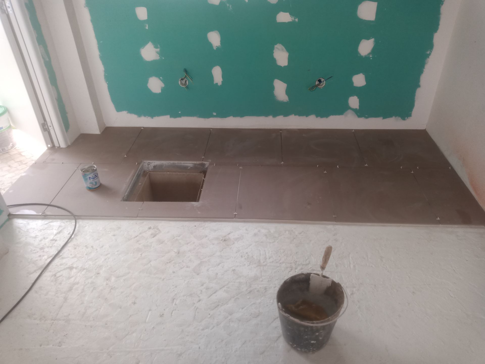 Canteiro de obras: piso de ladrilhos marrons com uma abertura quadrada. Parede verde com drywall, piso branco e balde com ferramentas.