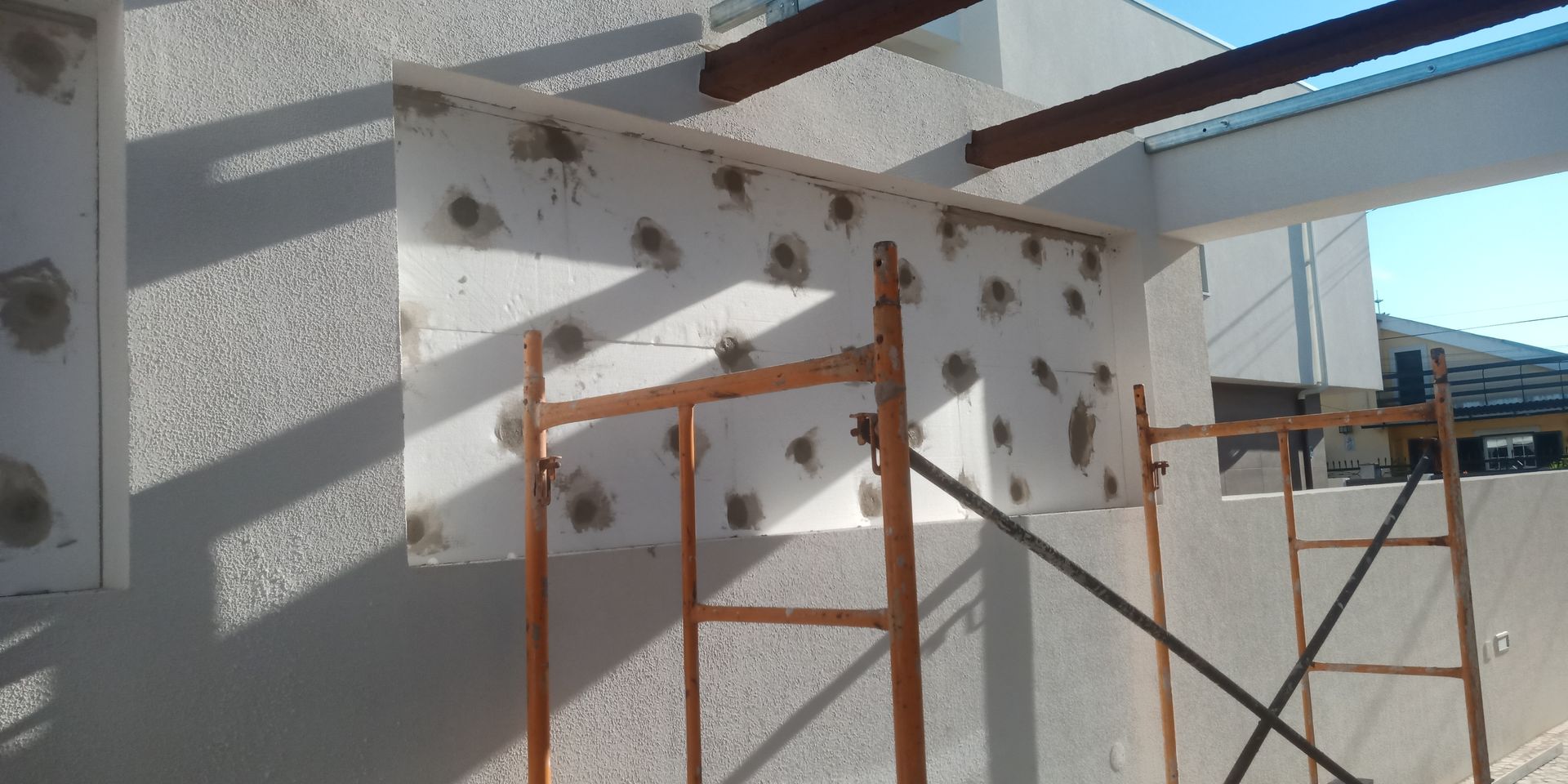 Canteiro de obras: placa de isolamento de espuma branca com manchas escuras fixada a um edifício, sustentada por andaimes laranja.