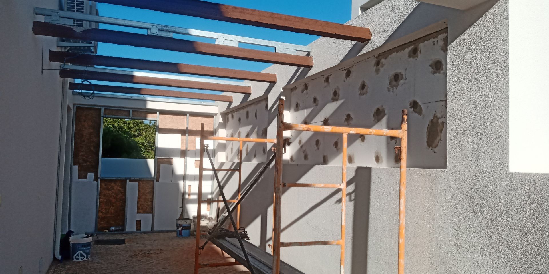 Canteiro de obras com vigas de madeira e andaimes encostados em uma parede texturizada, sob luz solar intensa.