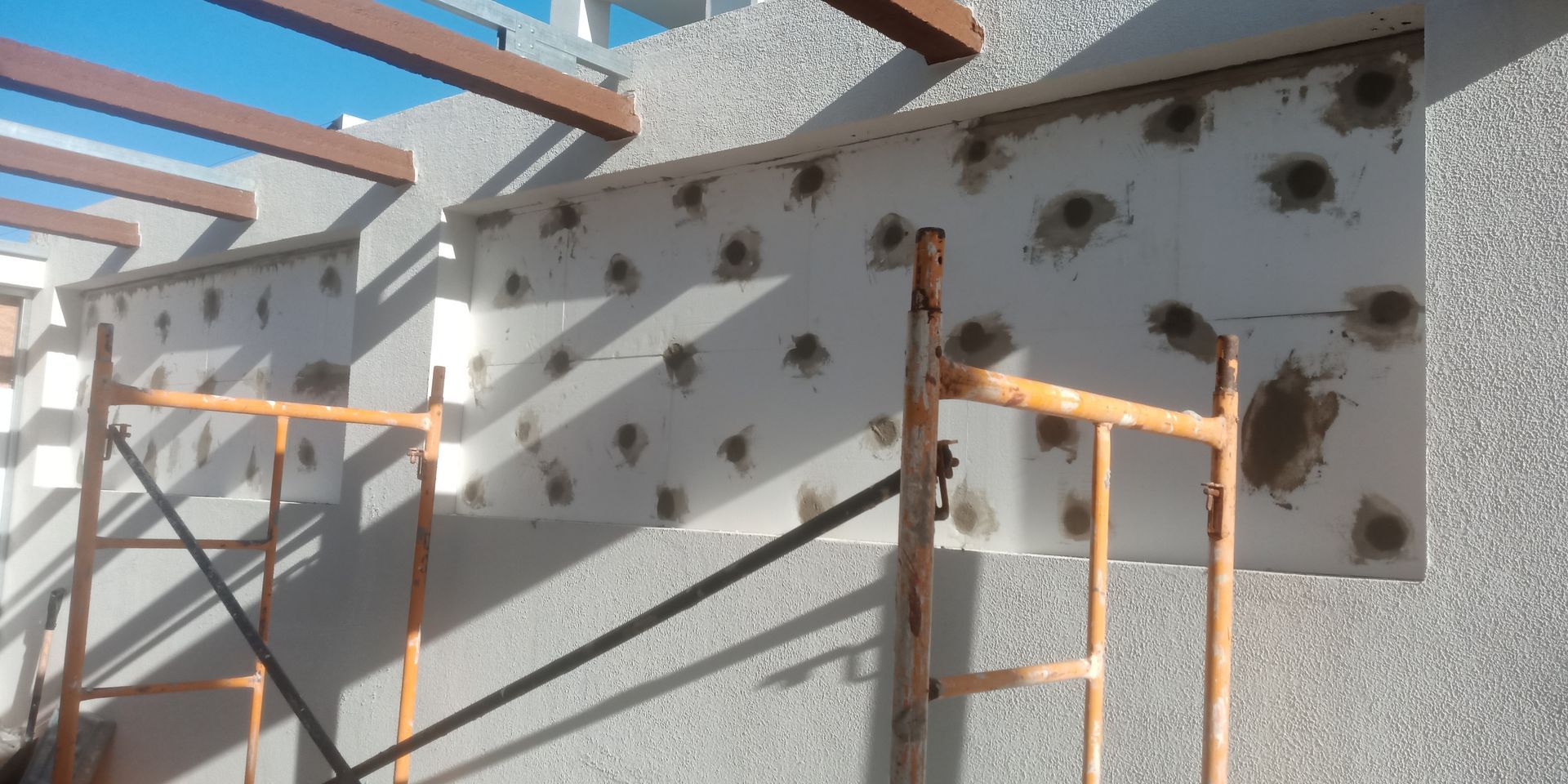 Canteiro de obras: andaimes junto a uma parede com buracos visíveis, possivelmente para isolamento ou reparo.