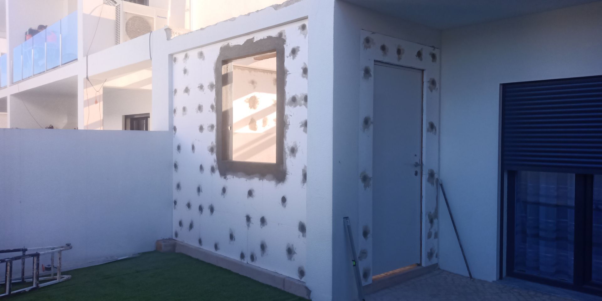 Edifício externo com janela e porta, cercado por detalhes decorativos. Persianas azuis. Grama artificial verde.