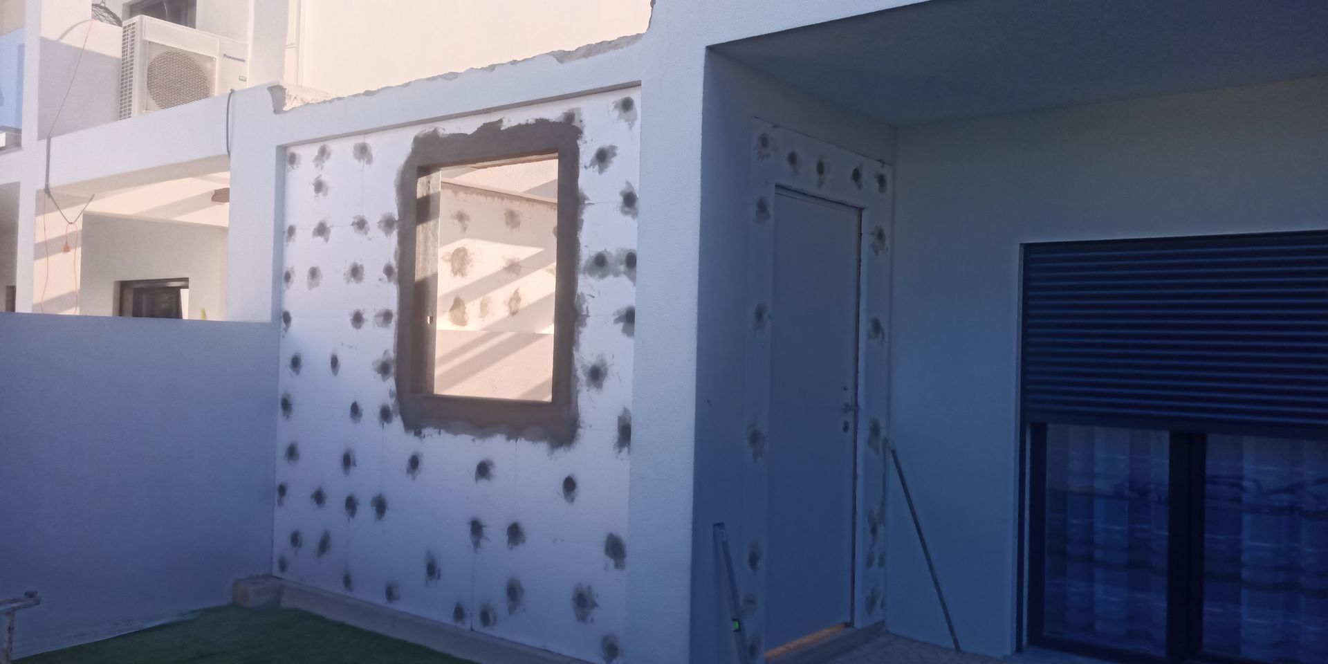 Parede externa com janela e porta, revestida com isolamento e pontos adesivos, em construção.