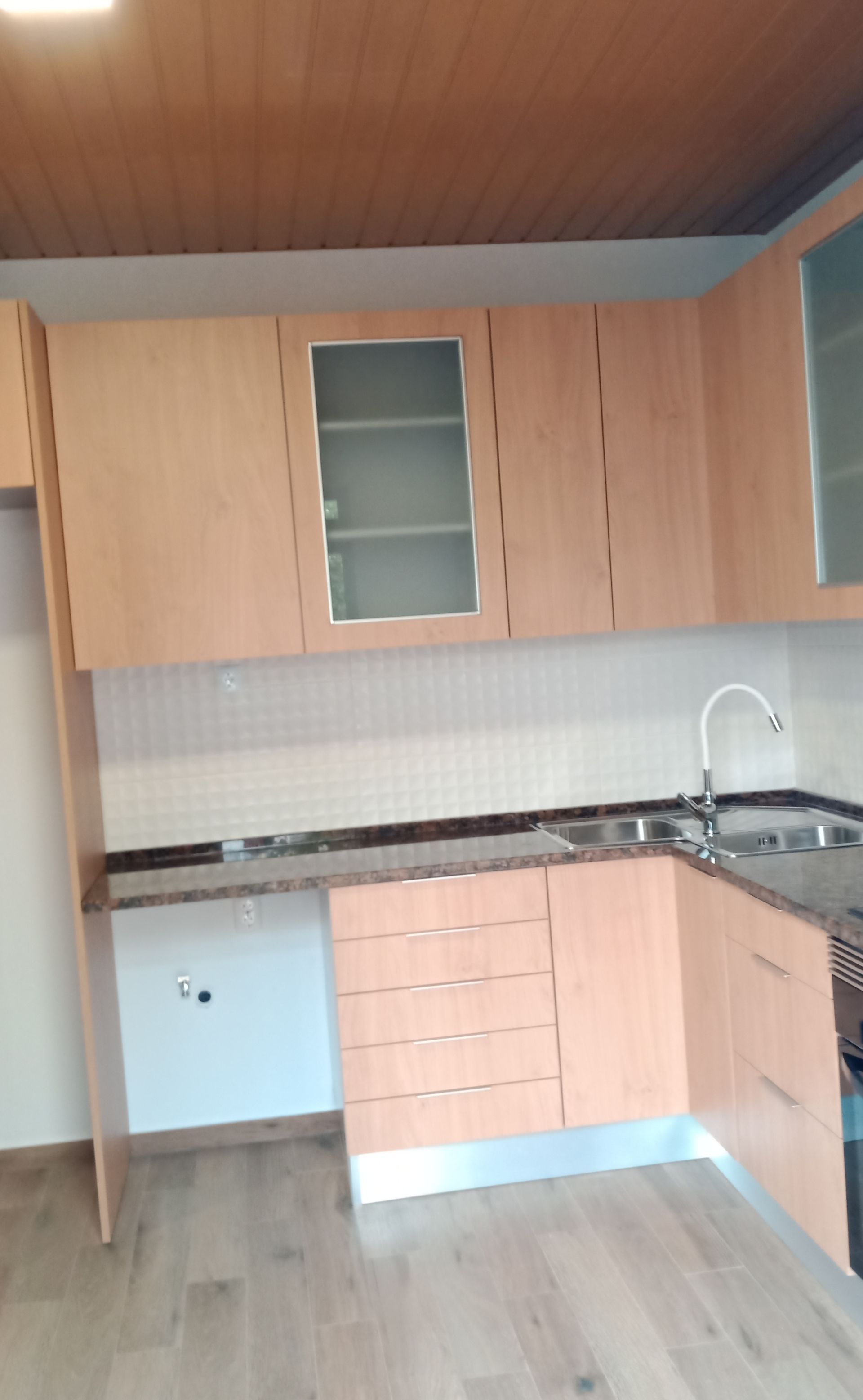 Armários de cozinha em madeira clara com portas de vidro na parte superior, bancada e pia de aço inoxidável.