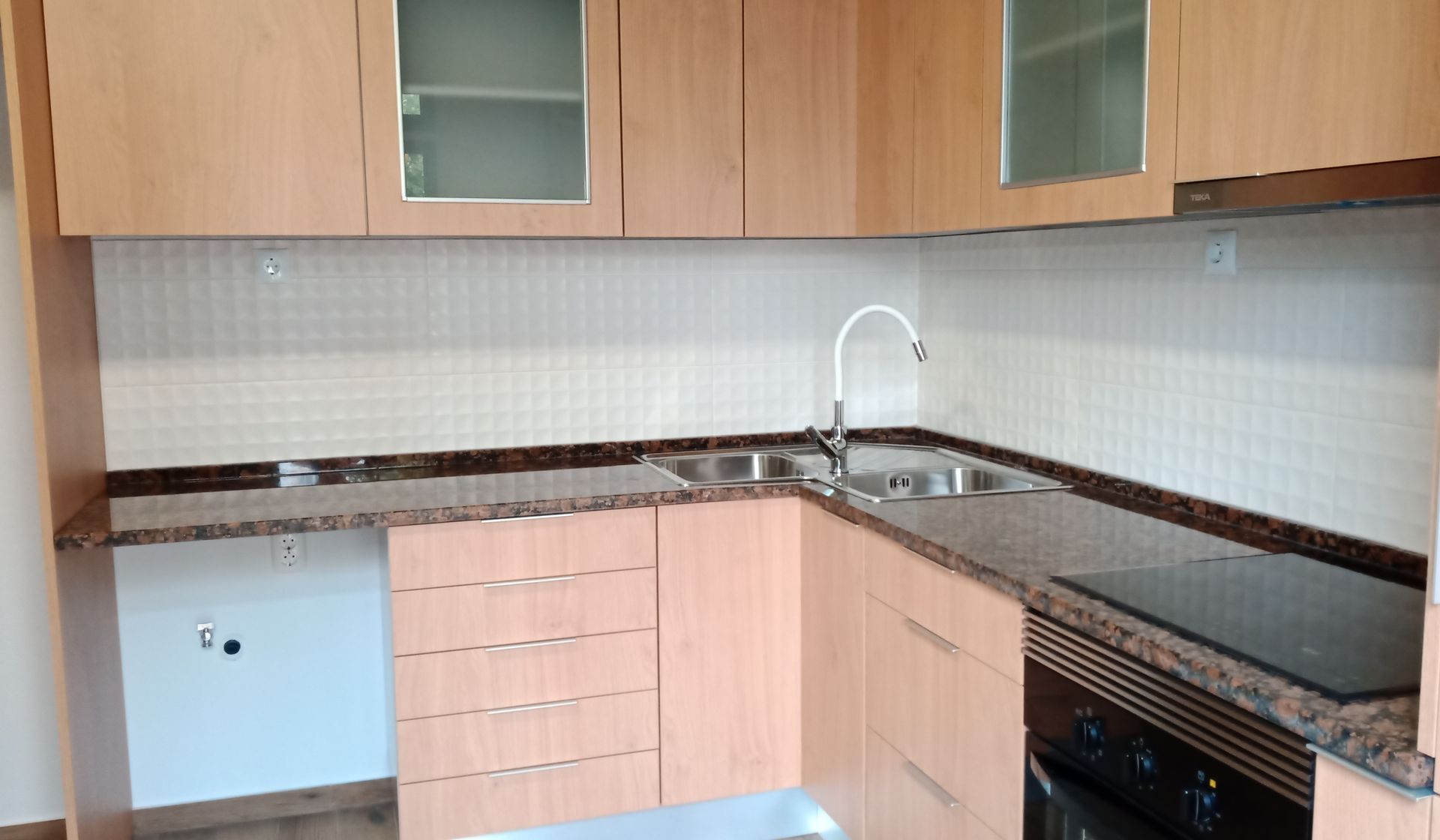 Armários e bancadas de cozinha em madeira clara com pia, fogão e painel de proteção contra respingos.