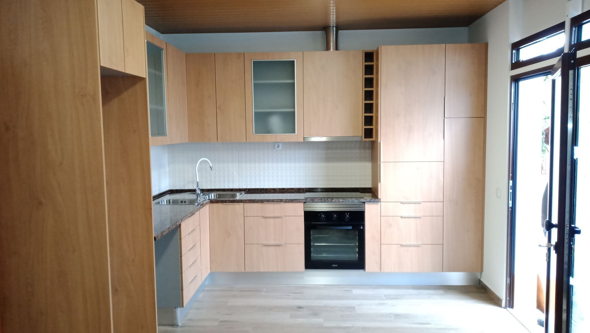 Cozinha moderna em madeira clara com armários, fogão, pia e porta aberta à direita.