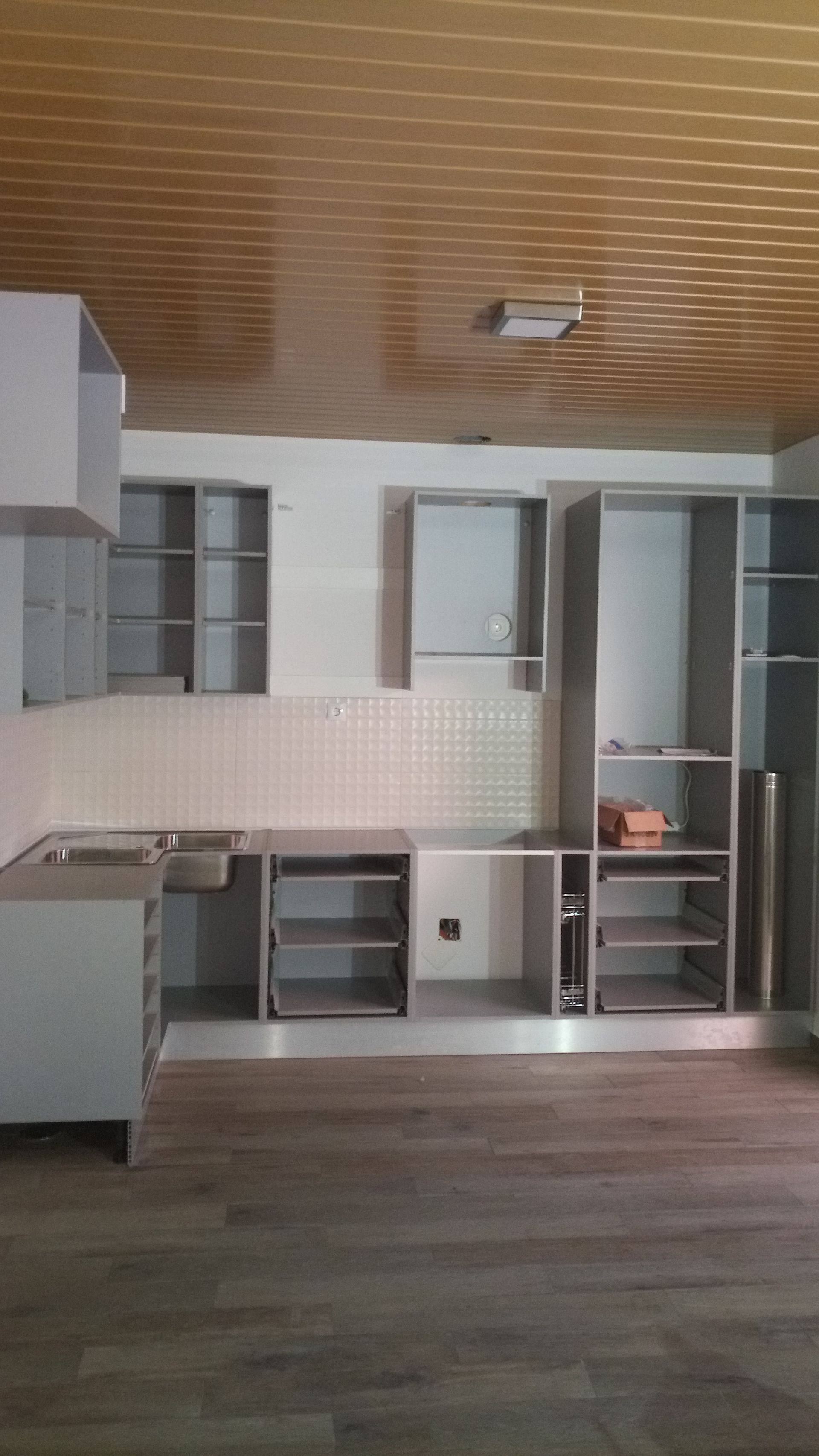 Armários de cozinha sendo instalados; cinza claro com portas e prateleiras abertas, contra backsplash branco e piso com aparência de madeira.
