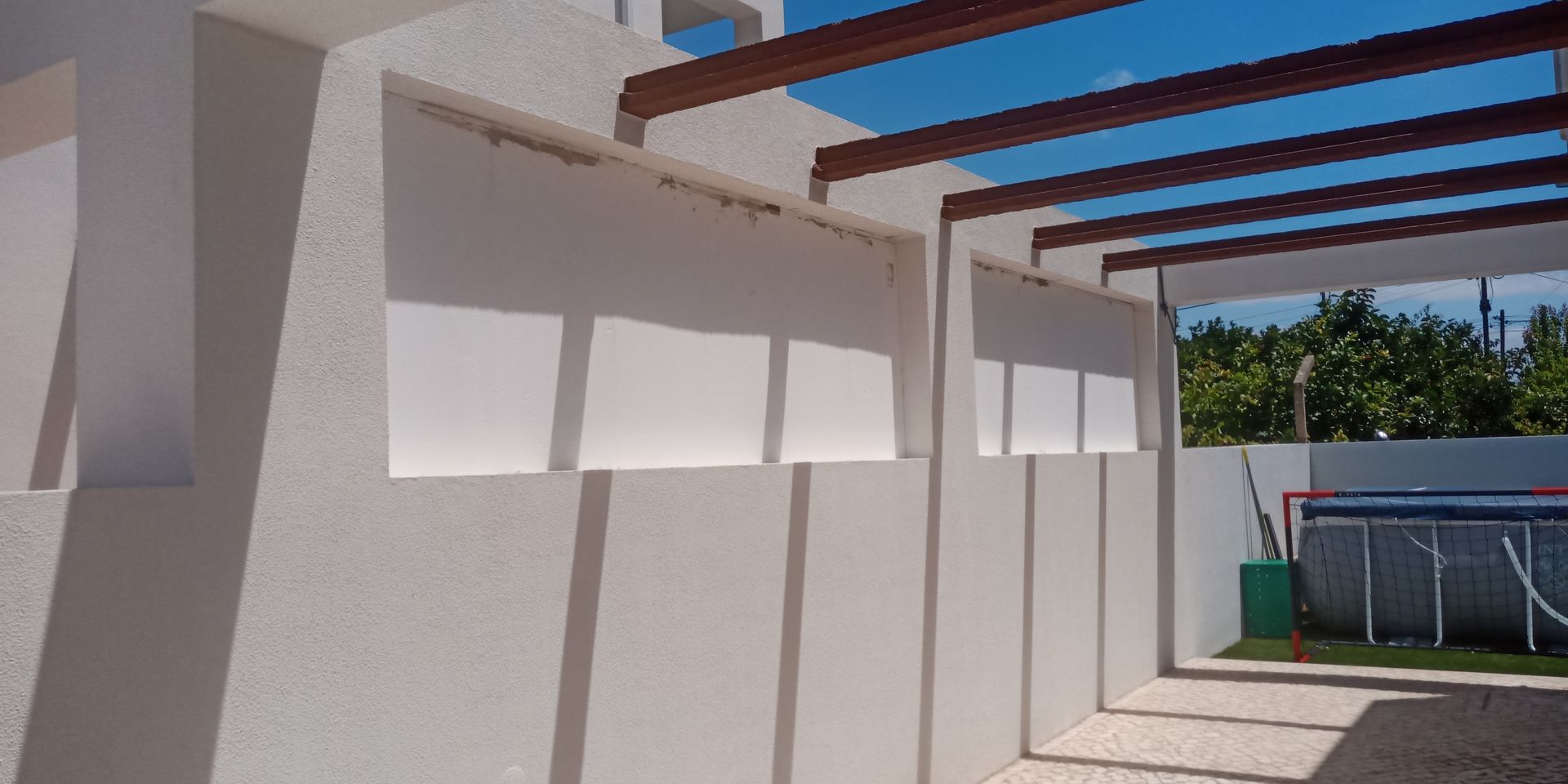 Exterior branco com telhado ripado, projetando sombras, e uma piscina ao fundo.