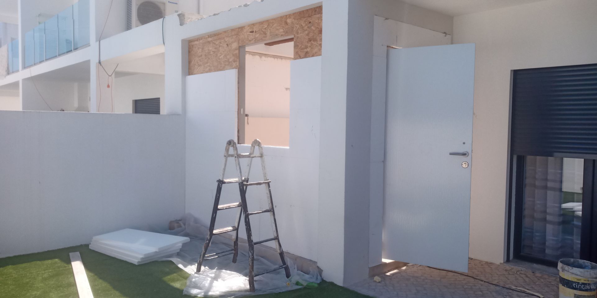 Vista exterior de um edifício em construção. Paredes brancas com uma porta aberta, uma escada e grama sintética.