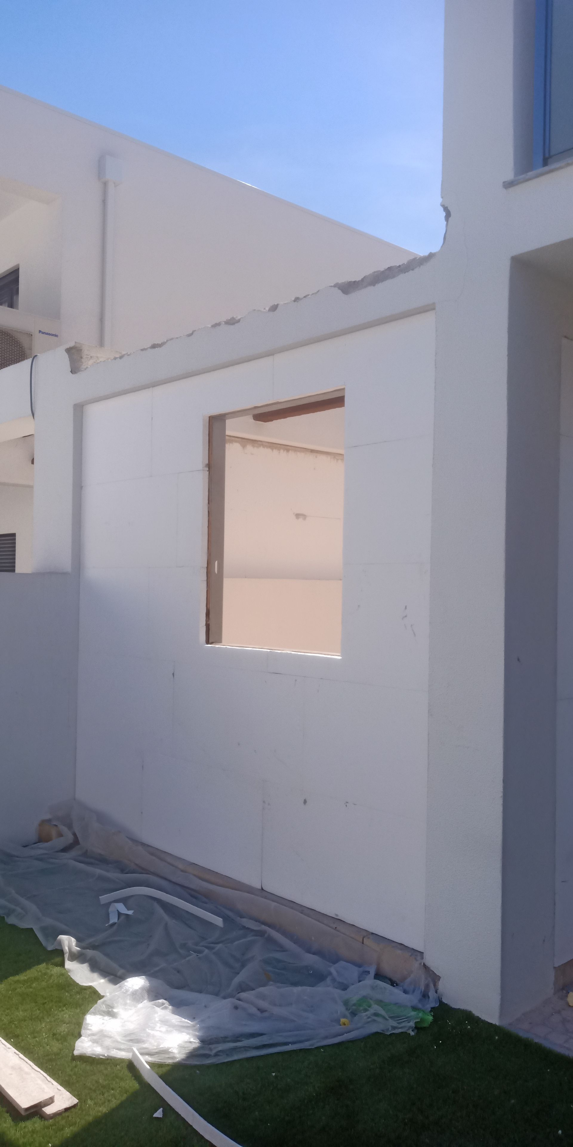 Vista exterior de um edifício em construção, com paredes brancas, moldura de janela e grama verde.