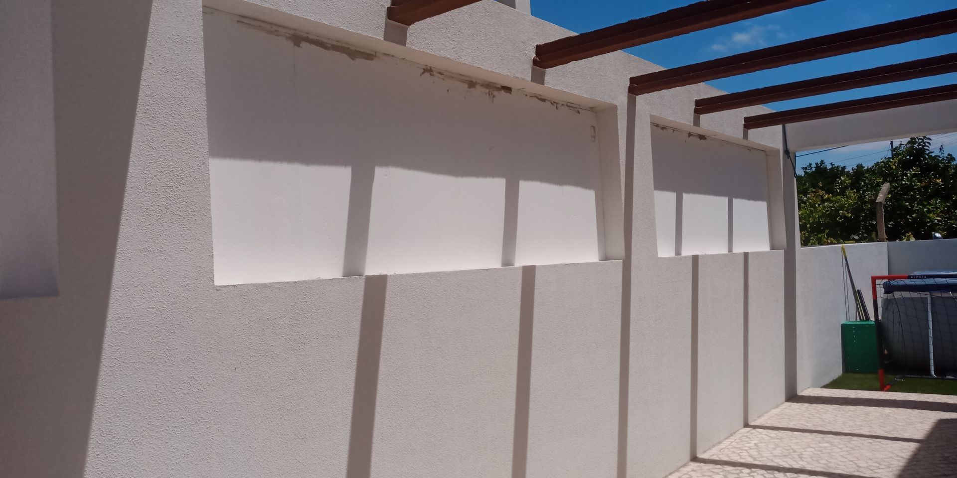 Persianas externas brancas em uma parede branca texturizada, sob uma pérgola de madeira, céu azul brilhante.