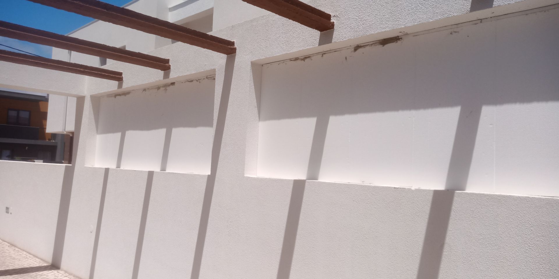 Prédio branco com aberturas horizontais e sombras projetadas por vigas no teto. Céu azul visível.