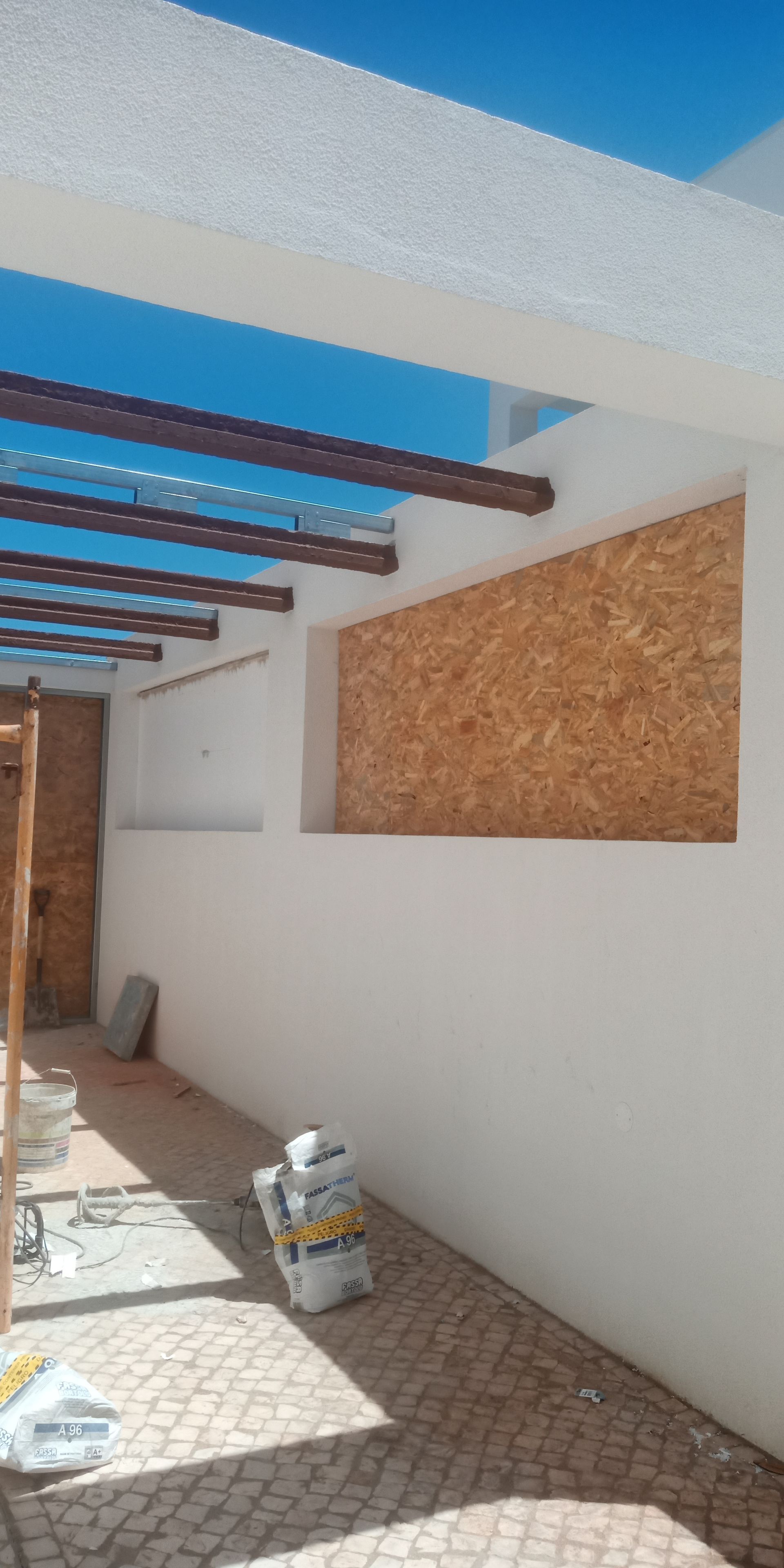 Área externa com paredes brancas, teto com vigas de madeira e janela fechada com tábuas de compensado marrom.