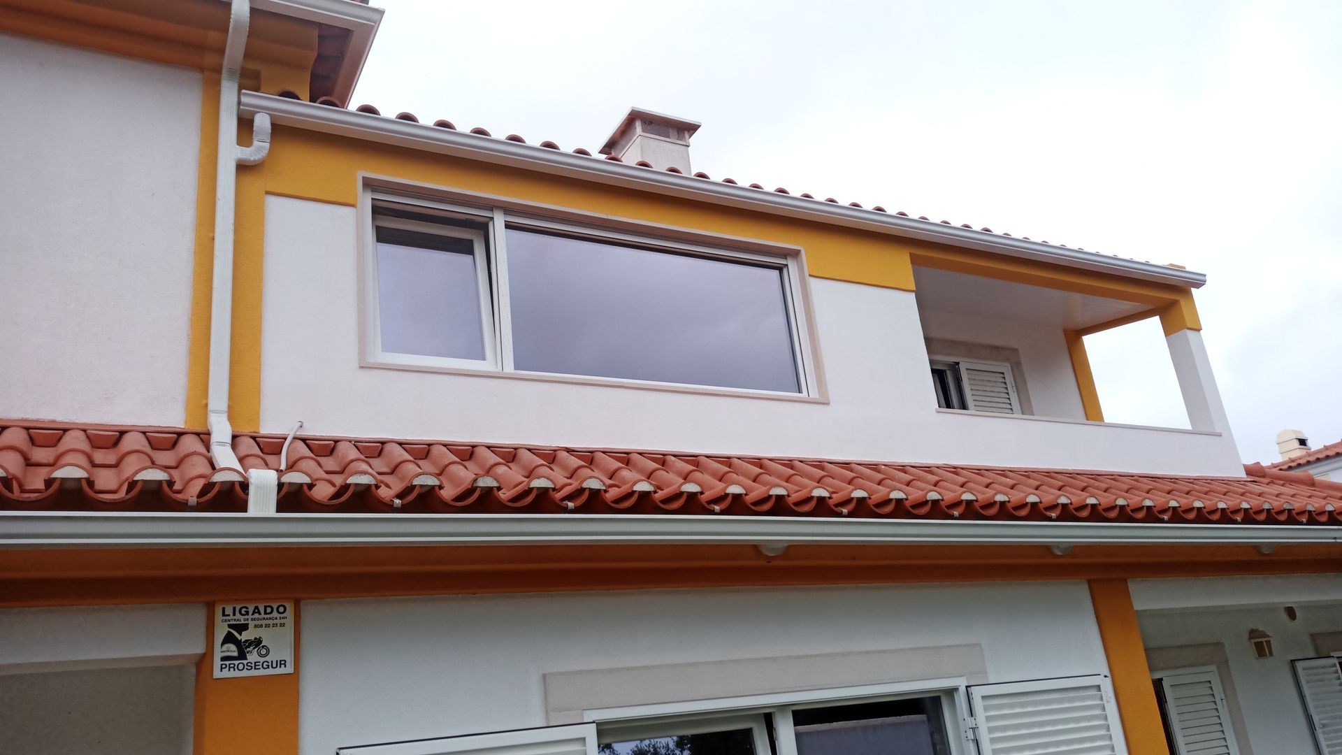 Casa de dois andares com telhas laranja, paredes brancas e detalhes em amarelo sob um céu nublado.