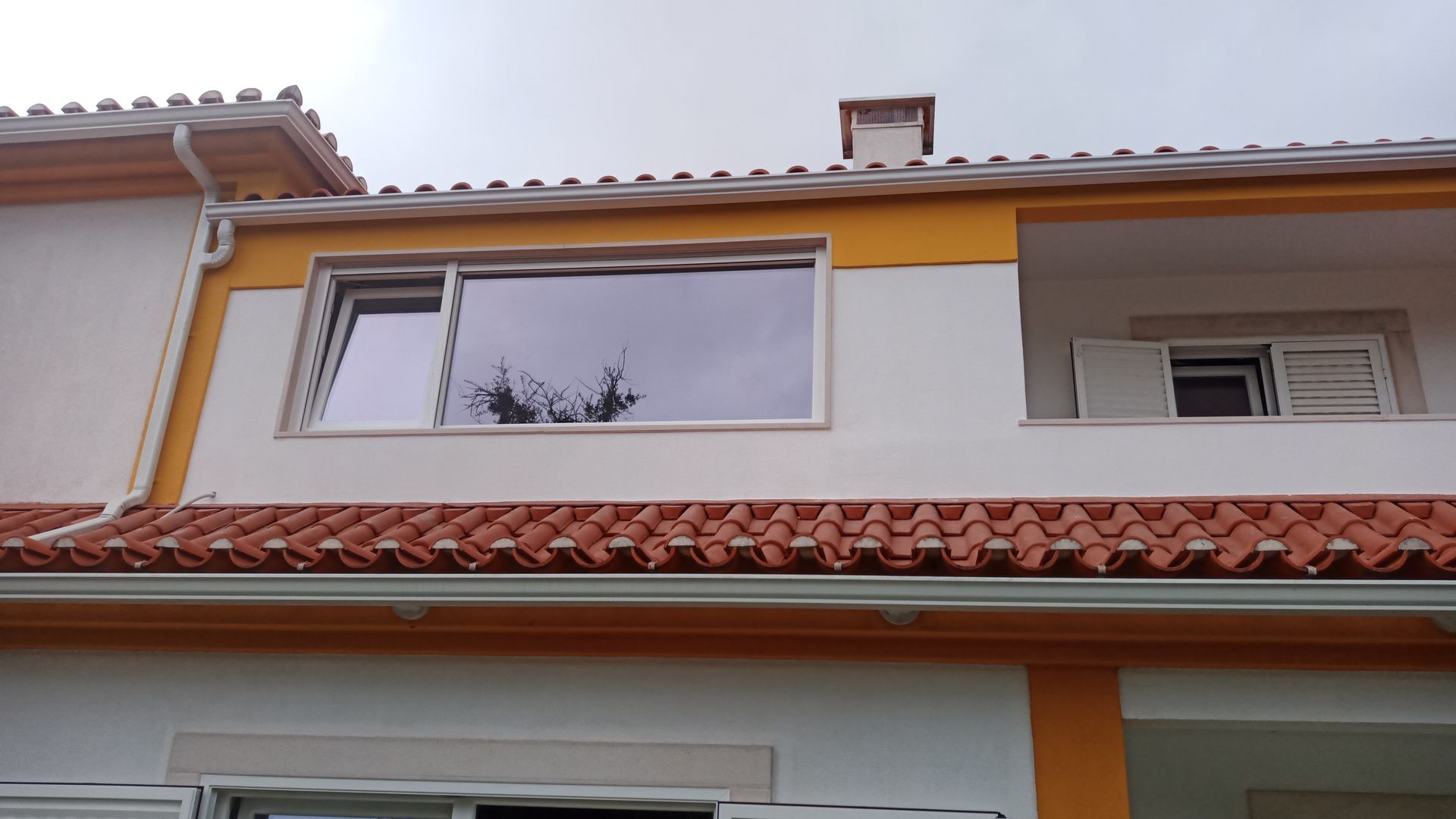 Prédio de dois andares com paredes brancas, detalhes em laranja e telhado de telha vermelha. Uma grande janela reflete as árvores.