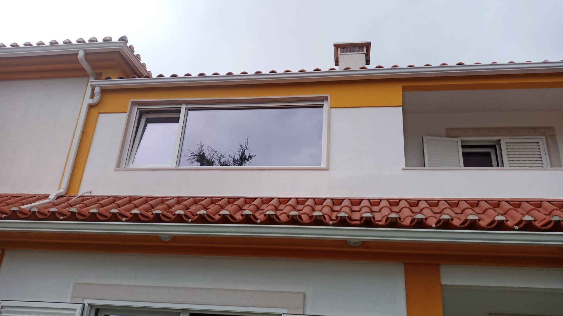 Casa de dois andares com telhado de telha vermelha, paredes brancas e amarelas, uma janela que reflete as árvores e um céu nublado.