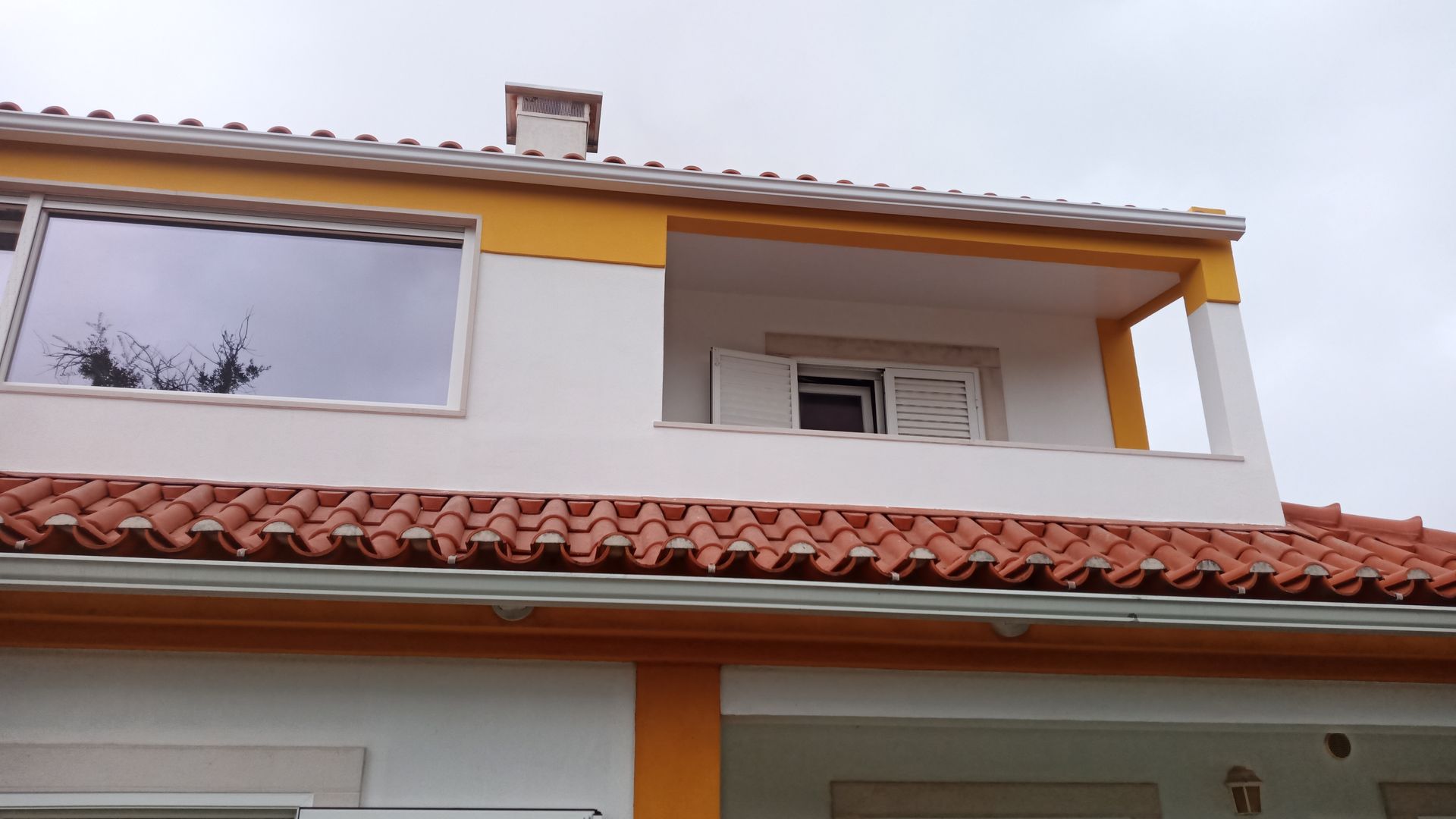 Casa branca de dois andares com detalhes em laranja, telhado de telha vermelha e varanda com venezianas abertas.