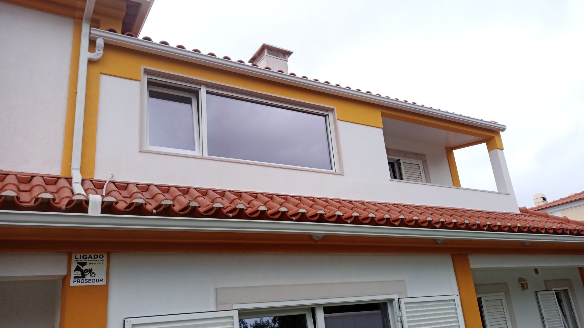 Casa de dois andares com detalhes em laranja, paredes brancas e telhado de telha vermelha; céu nublado acima.