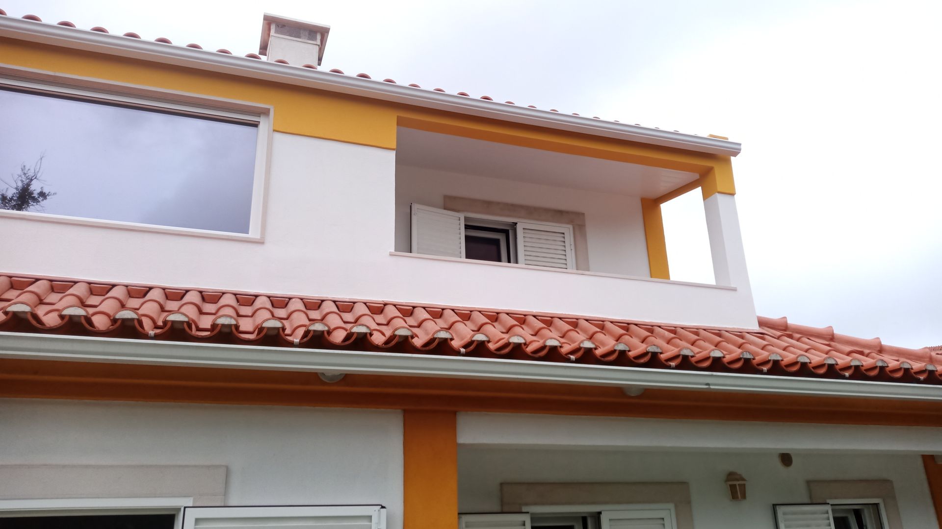 Casa de dois andares com paredes brancas, telhado laranja e detalhes em amarelo. Céu nublado.