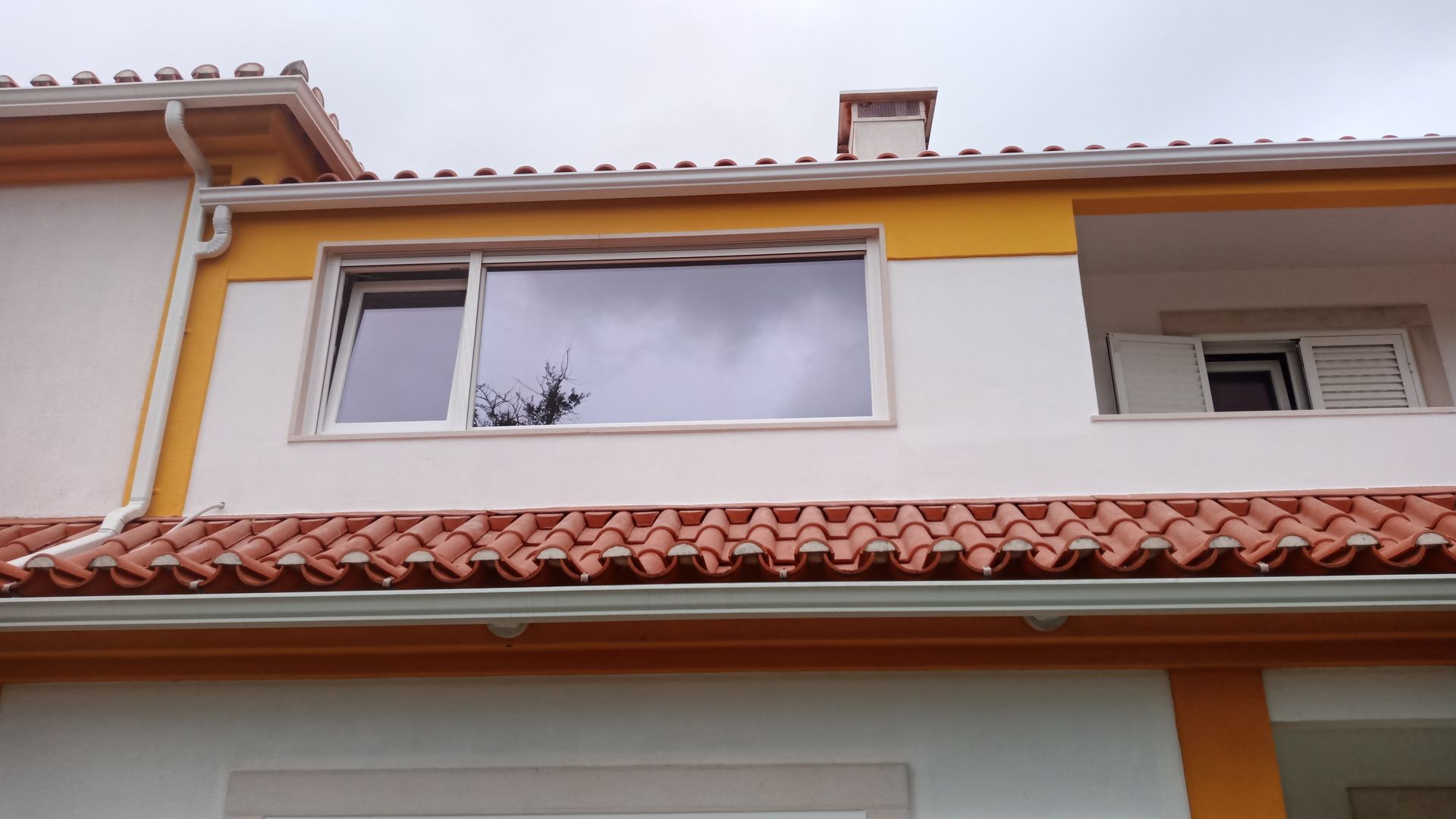 Prédio branco e amarelo com telhado de telhas vermelhas, janela e calha sob um céu nublado.