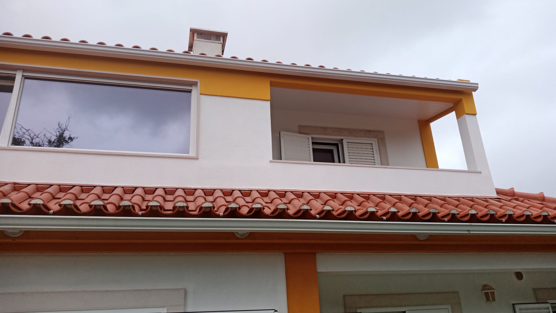 Prédio de dois andares com paredes brancas, detalhes em laranja e telhado de telha vermelha sob um céu nublado.