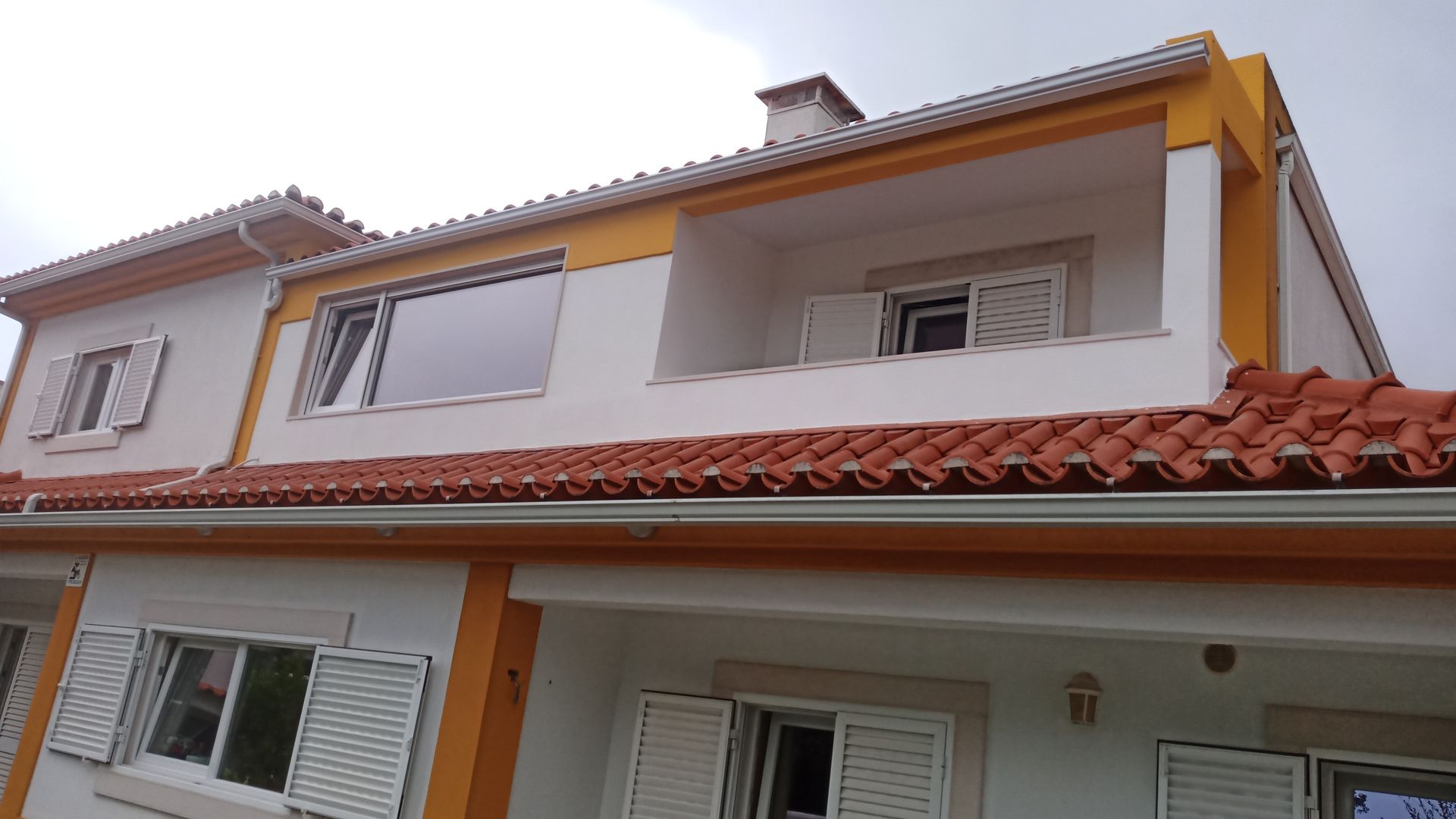 Casa branca de dois andares com detalhes em laranja e telhas de terracota, sob um céu nublado.