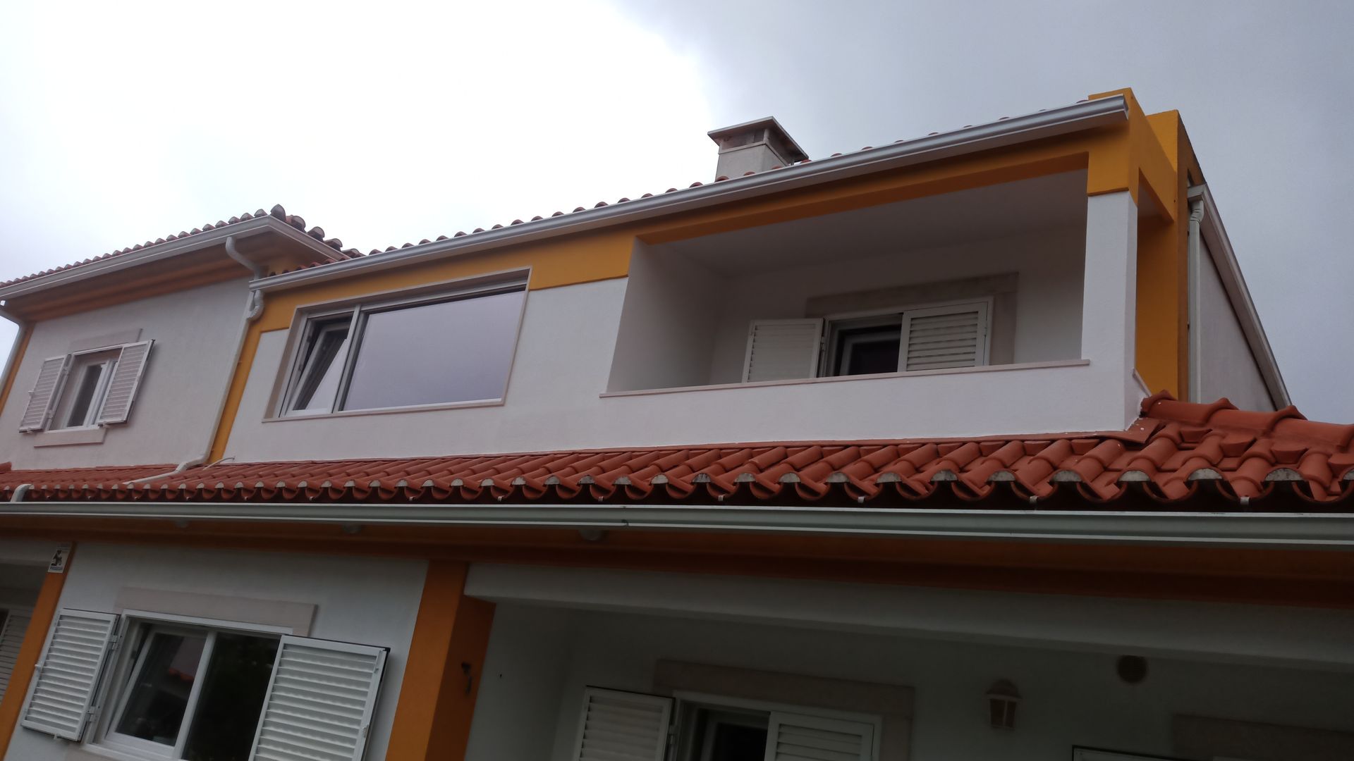 Casa de dois andares com telhas laranja, paredes brancas e detalhes em amarelo sob um céu nublado.